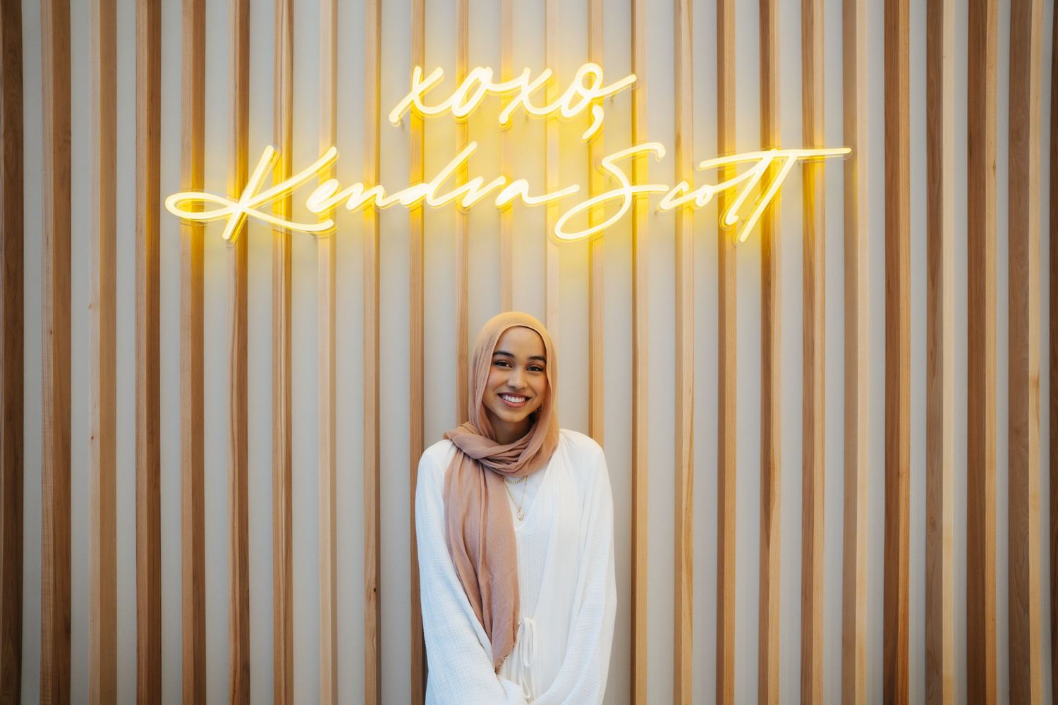 Woman in hijab smiling in front of XOXO Kendra Scott neon sign on wood wall.