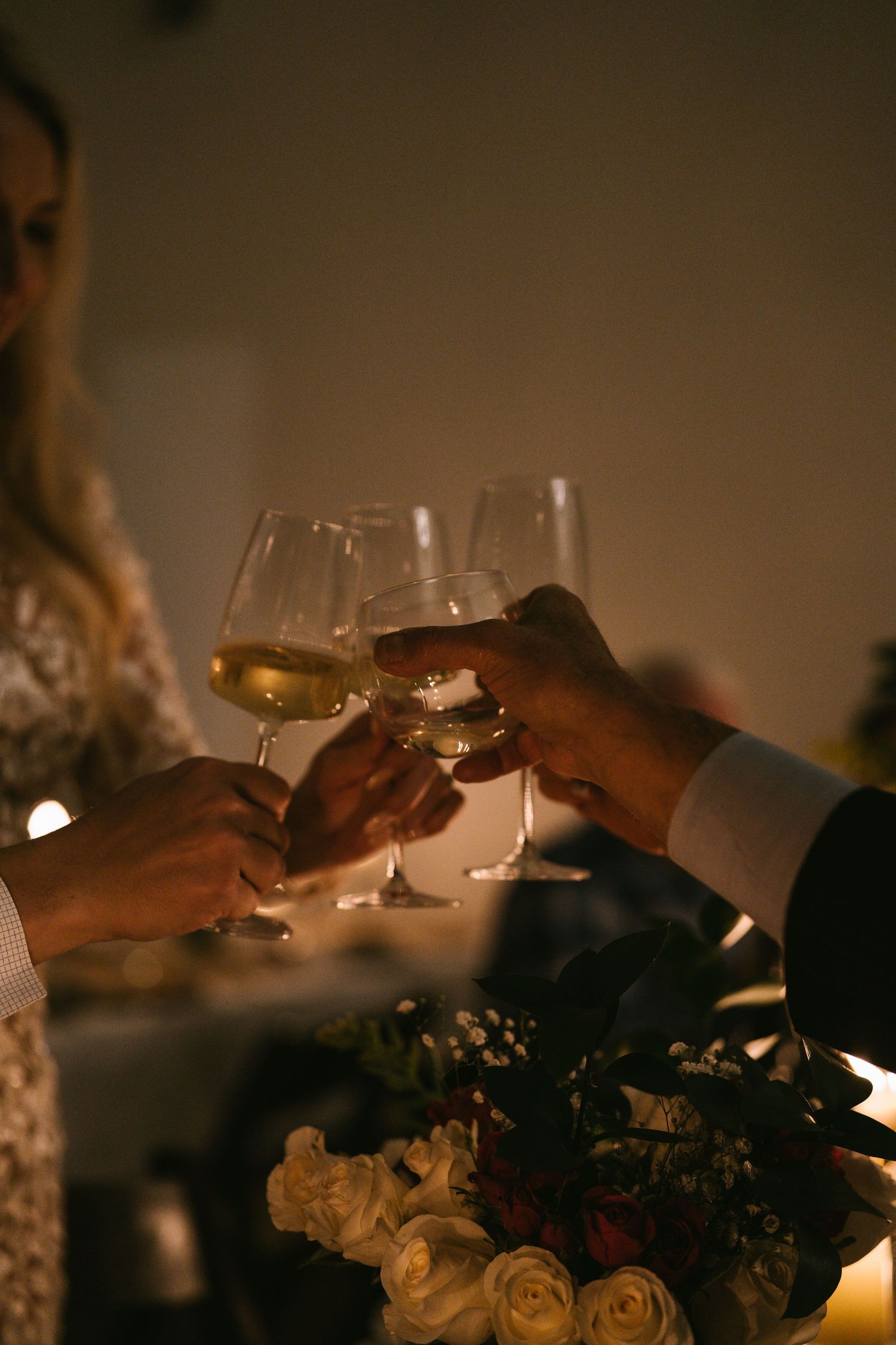 Hands clinking wine glasses over a floral centerpiece at a candlelit celebration.