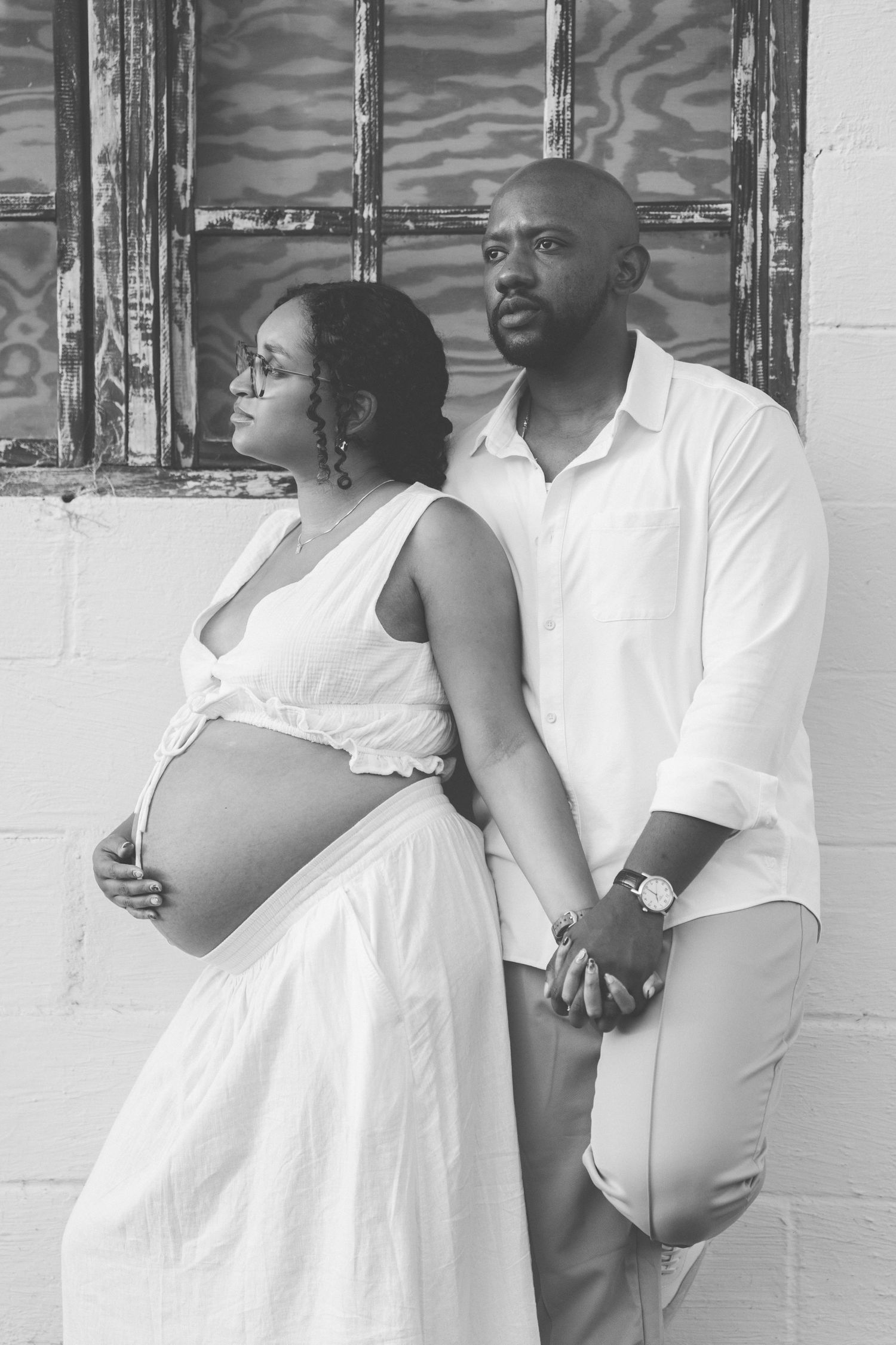Black and white maternity photo of couple posing against rustic wall.