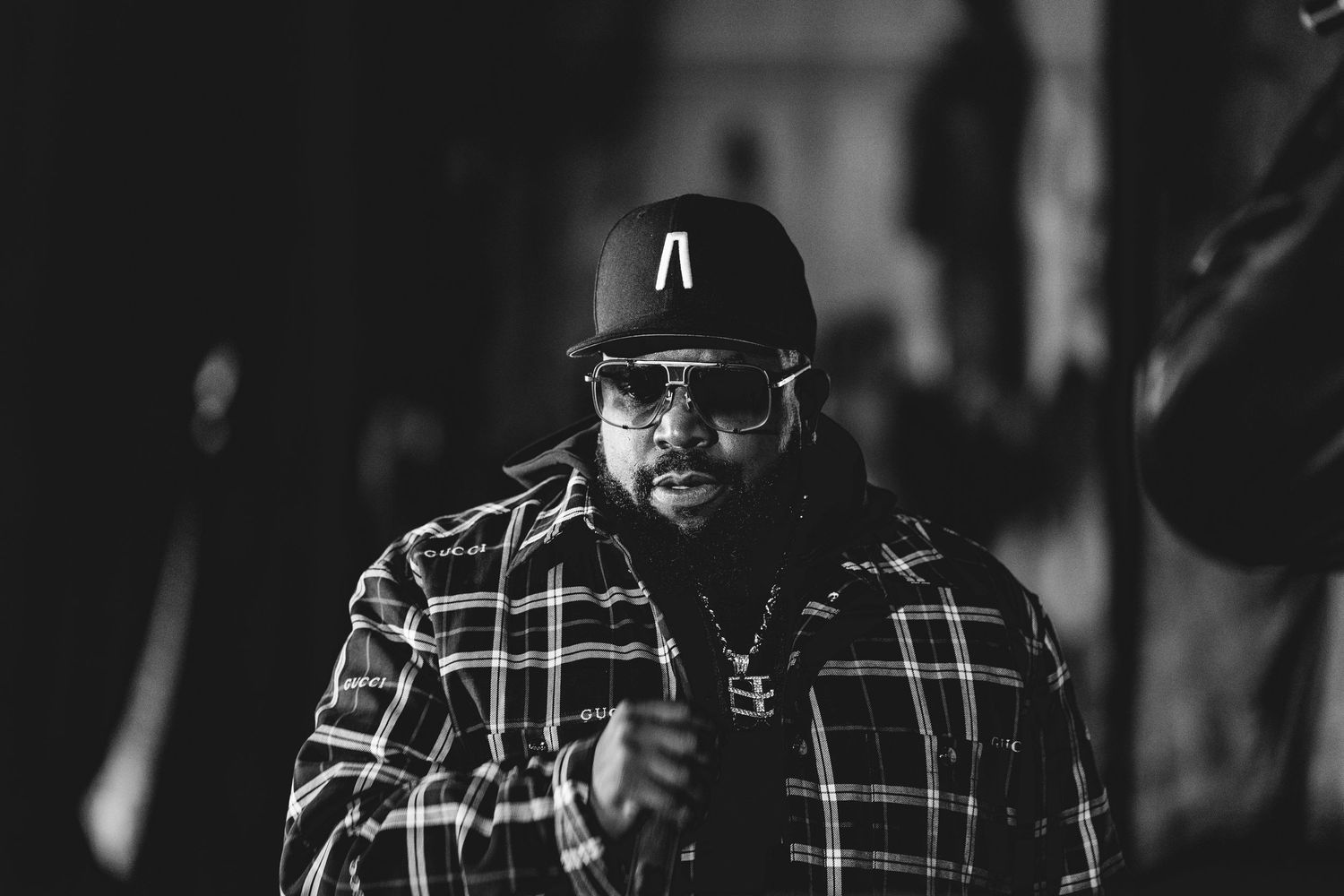 Black and white portrait of a man wearing a plaid shirt and snapback cap in a dimly lit setting.