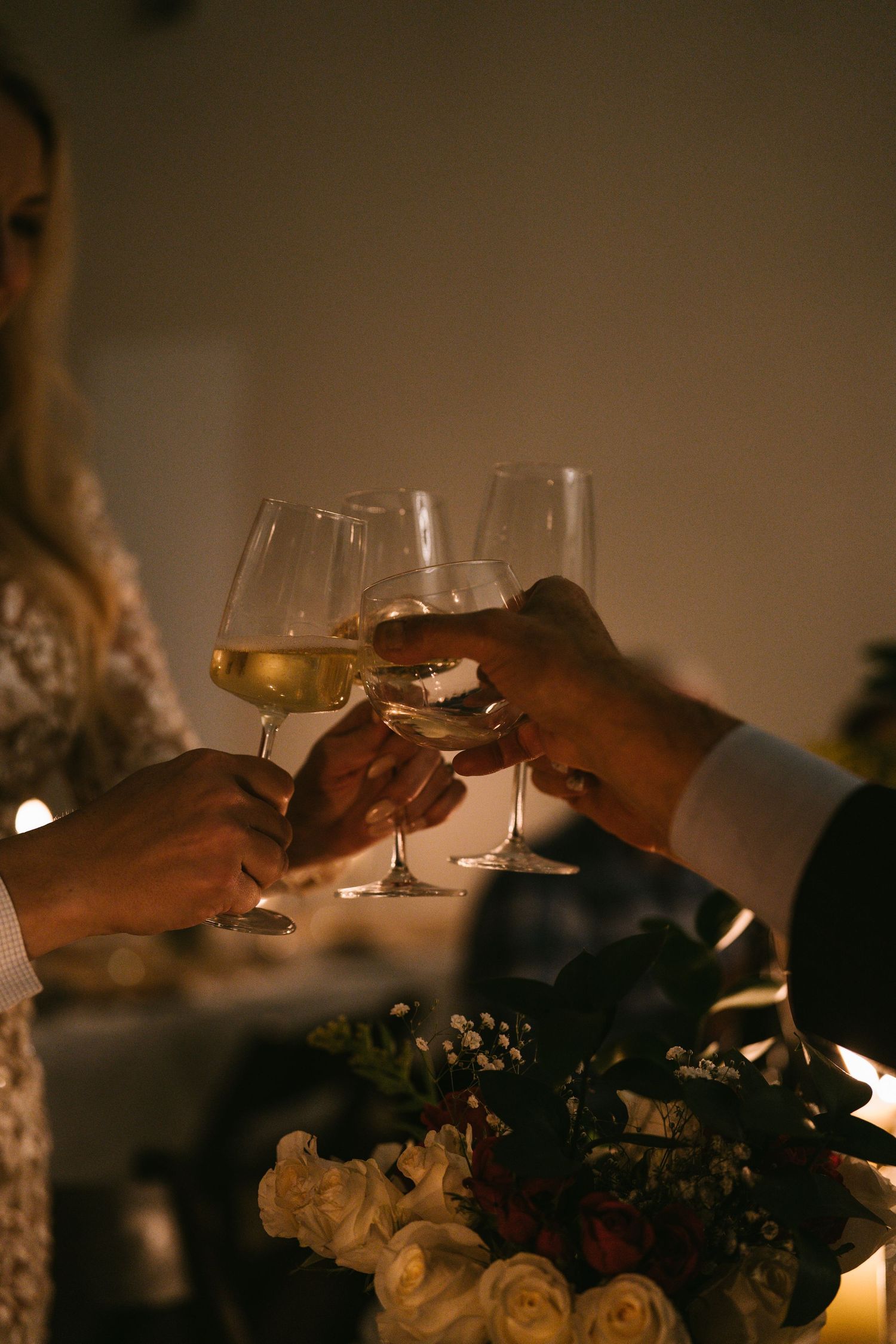 Hands clinking glasses of white wine over a floral centerpiece at a romantic candlelit wedding reception.