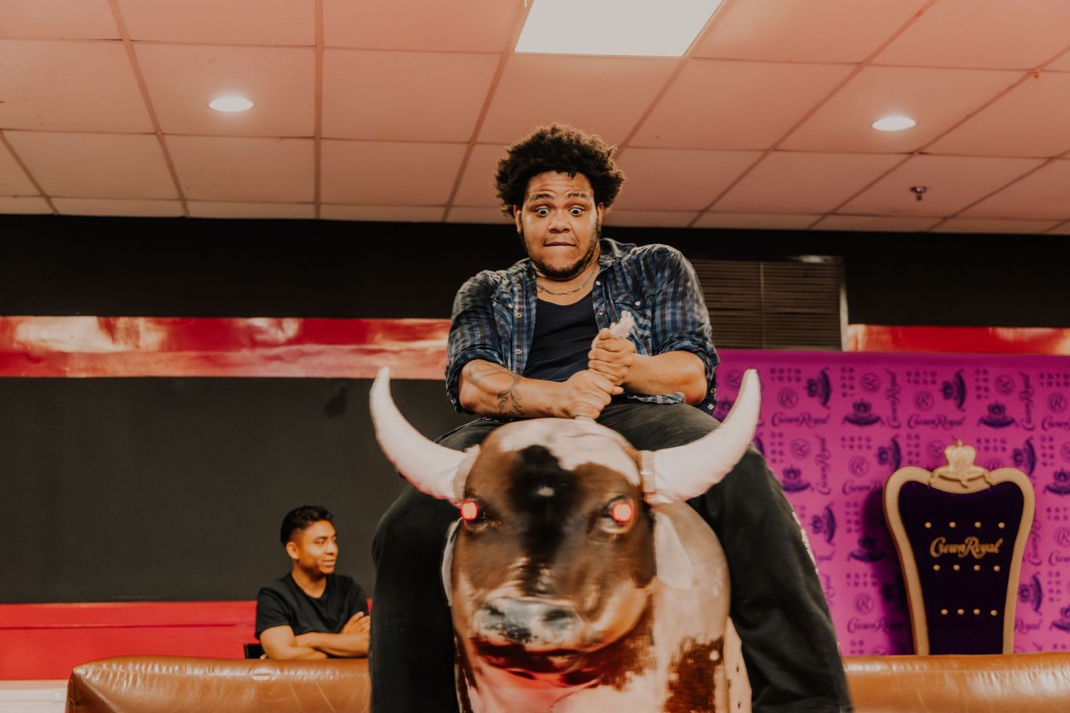 Young man smiles while riding a mechanical bull indoors.