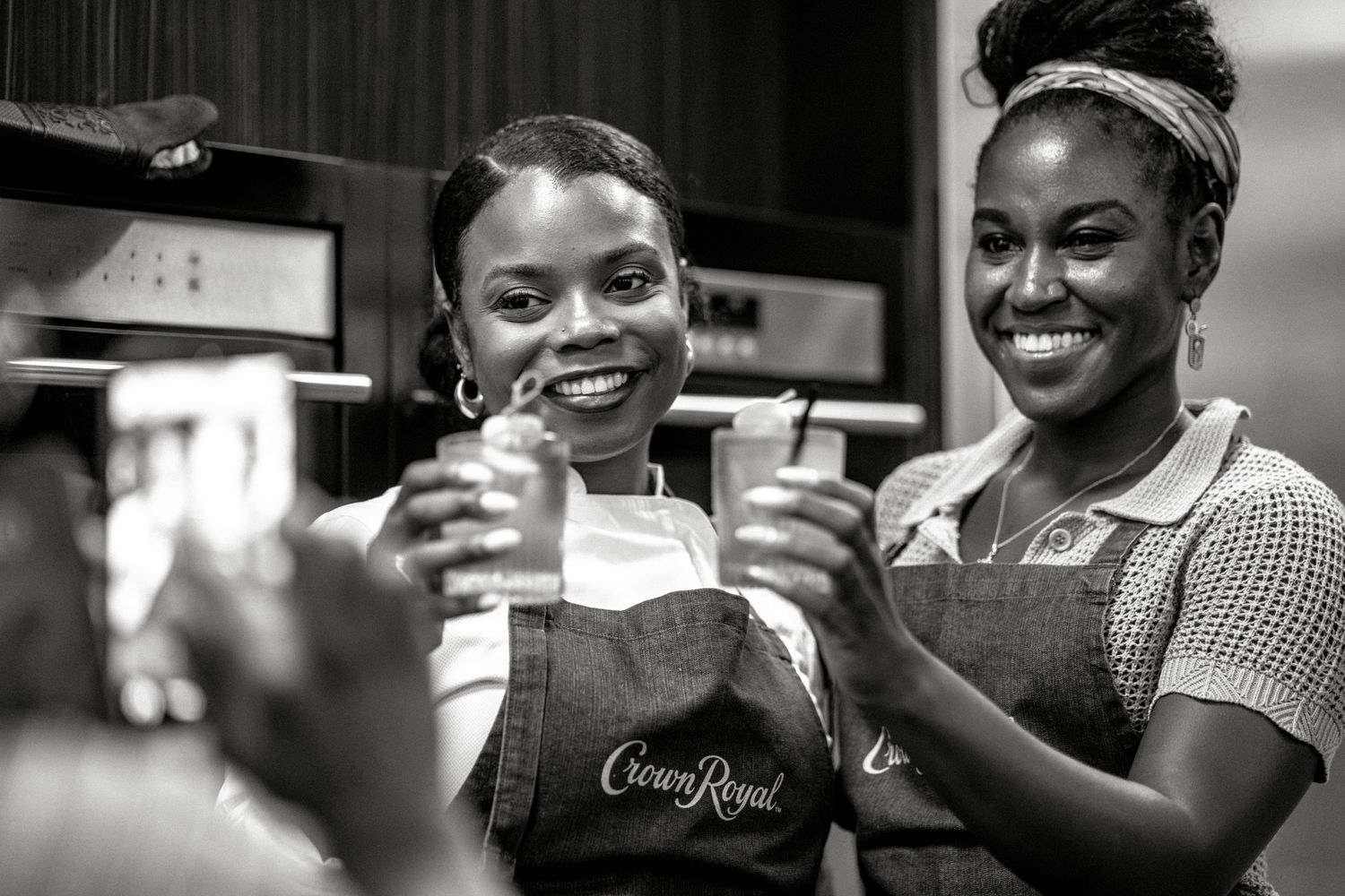 Two smiling women in Crown Royal aprons toast with cocktail glasses at a branded event.