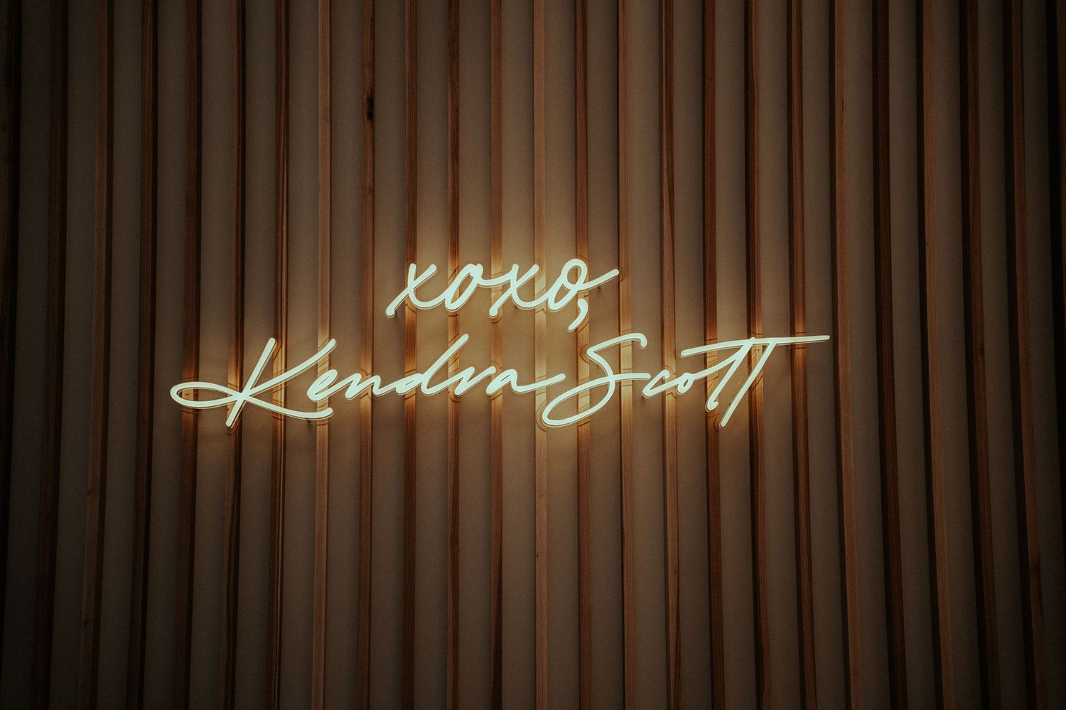 Neon sign reading XOXO Kendra Scott in cursive script on a wooden slatted wall backdrop.