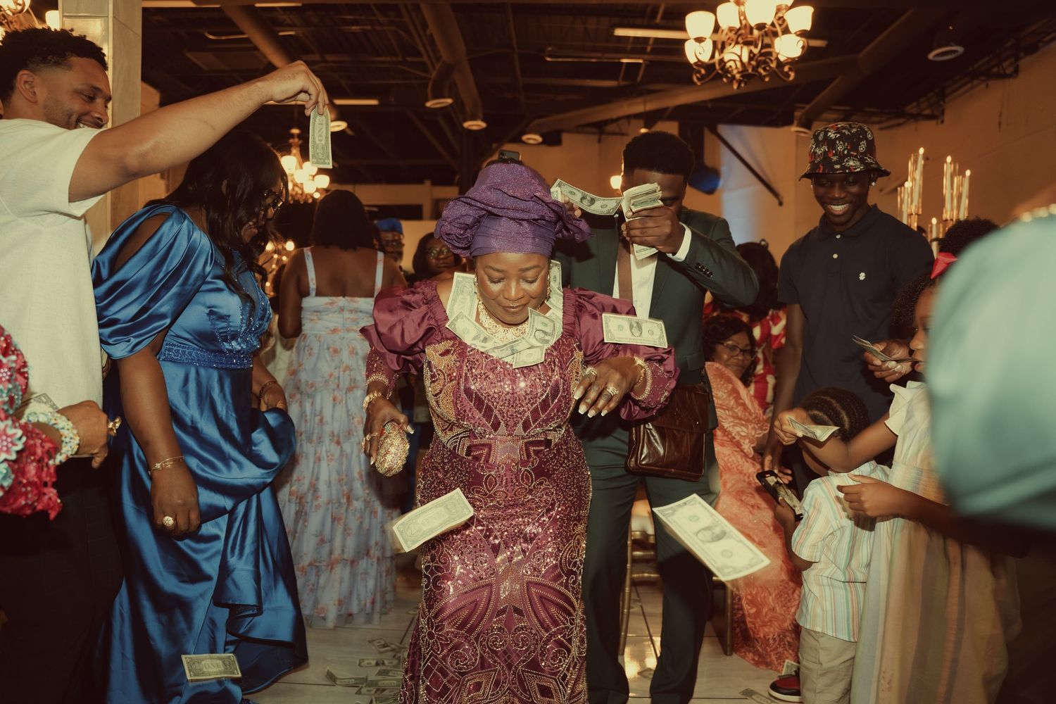 Woman in purple Nigerian traditional dress walking through a festive indoor celebration with guests.