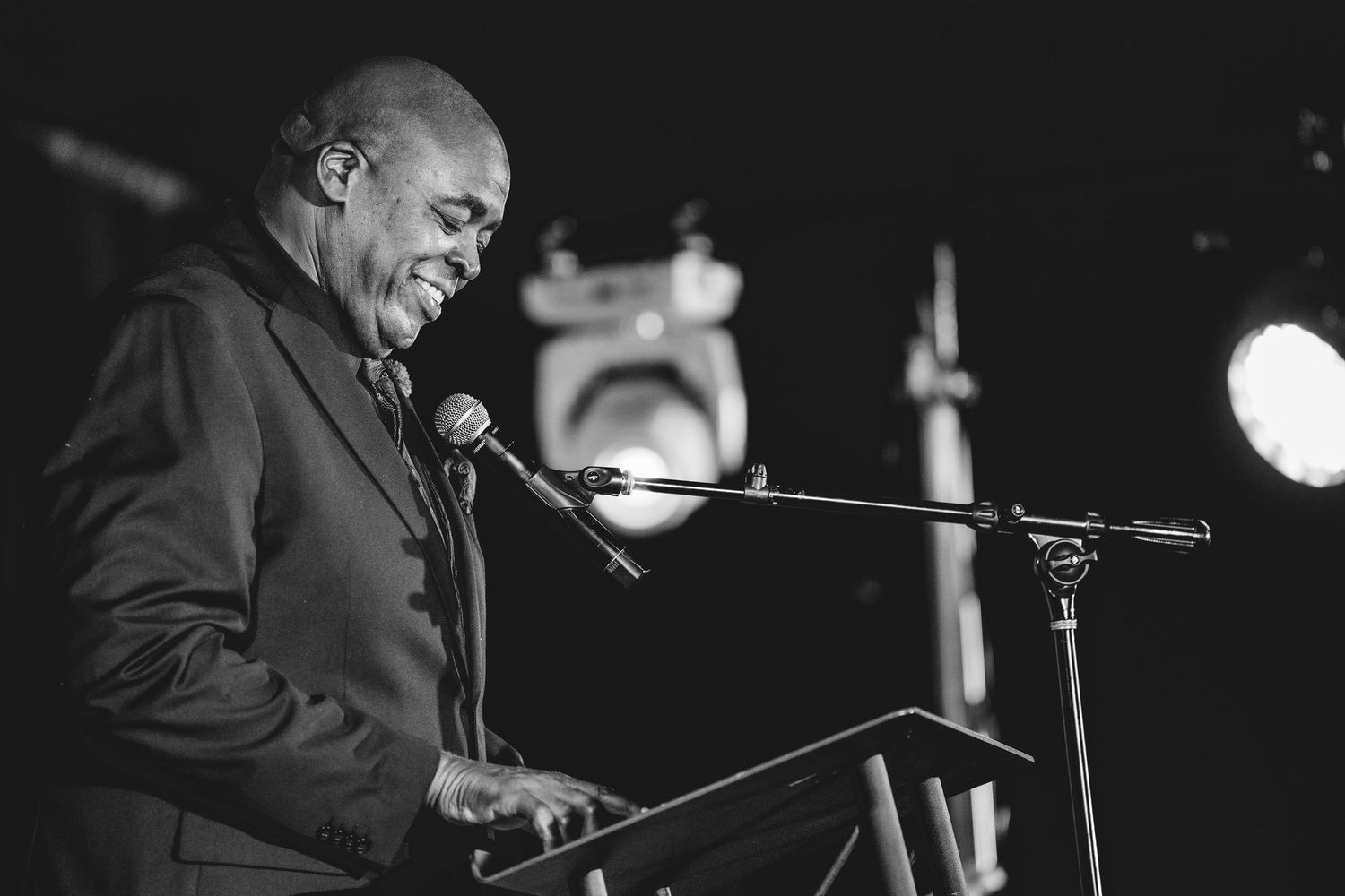Man in dark suit speaking at a podium with microphone under stage lighting in black and white photo.