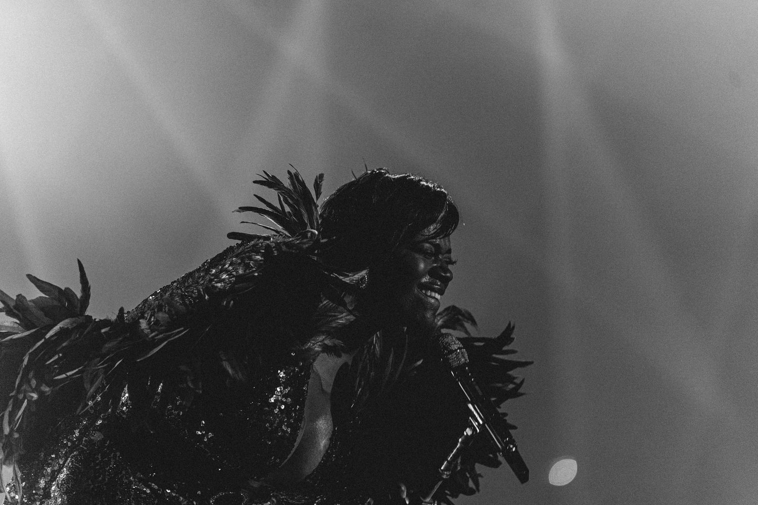 Black and white concert photo of performer in elaborate feathered outfit singing on a dramatic lit stage.