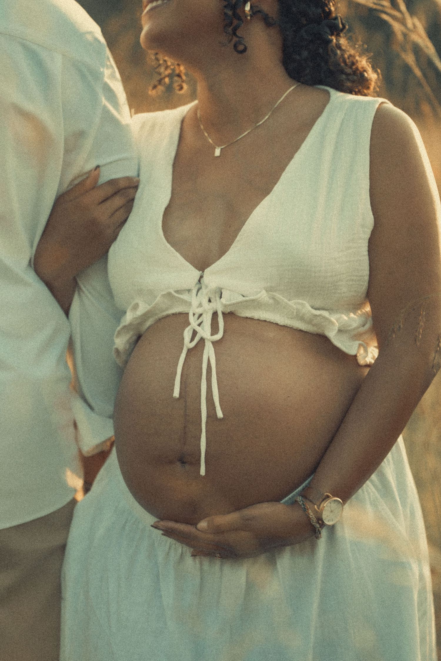 Pregnant woman in white dress holds baby bump outdoors in golden light.