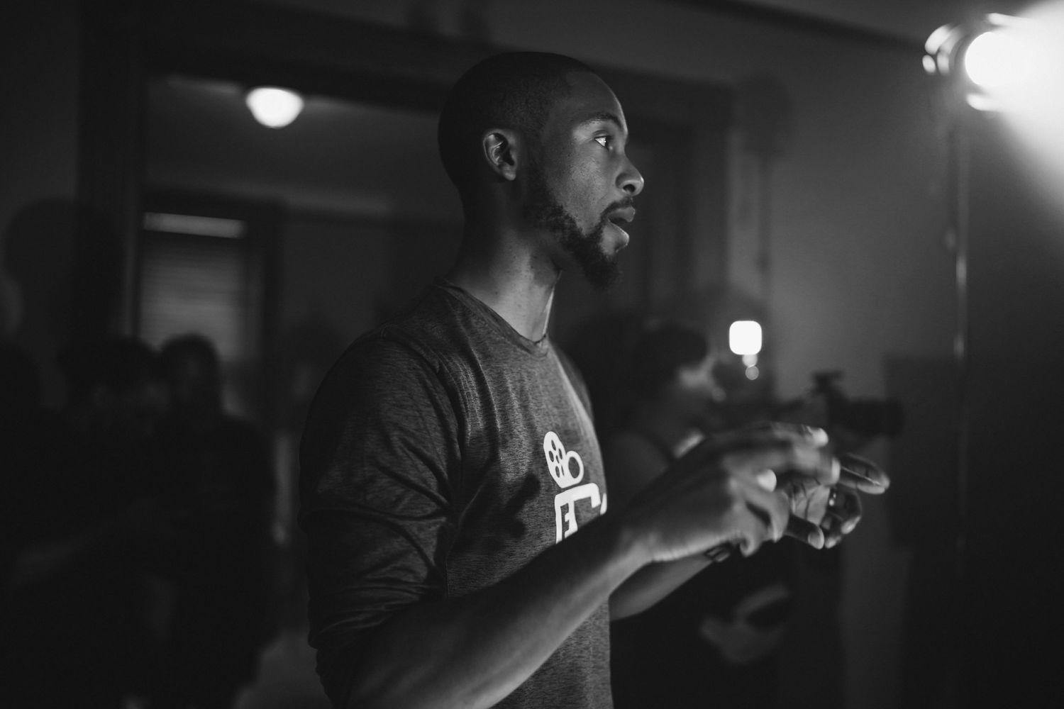 Black and white photo of a man gesturing with his hands under a studio light in a dark room.
