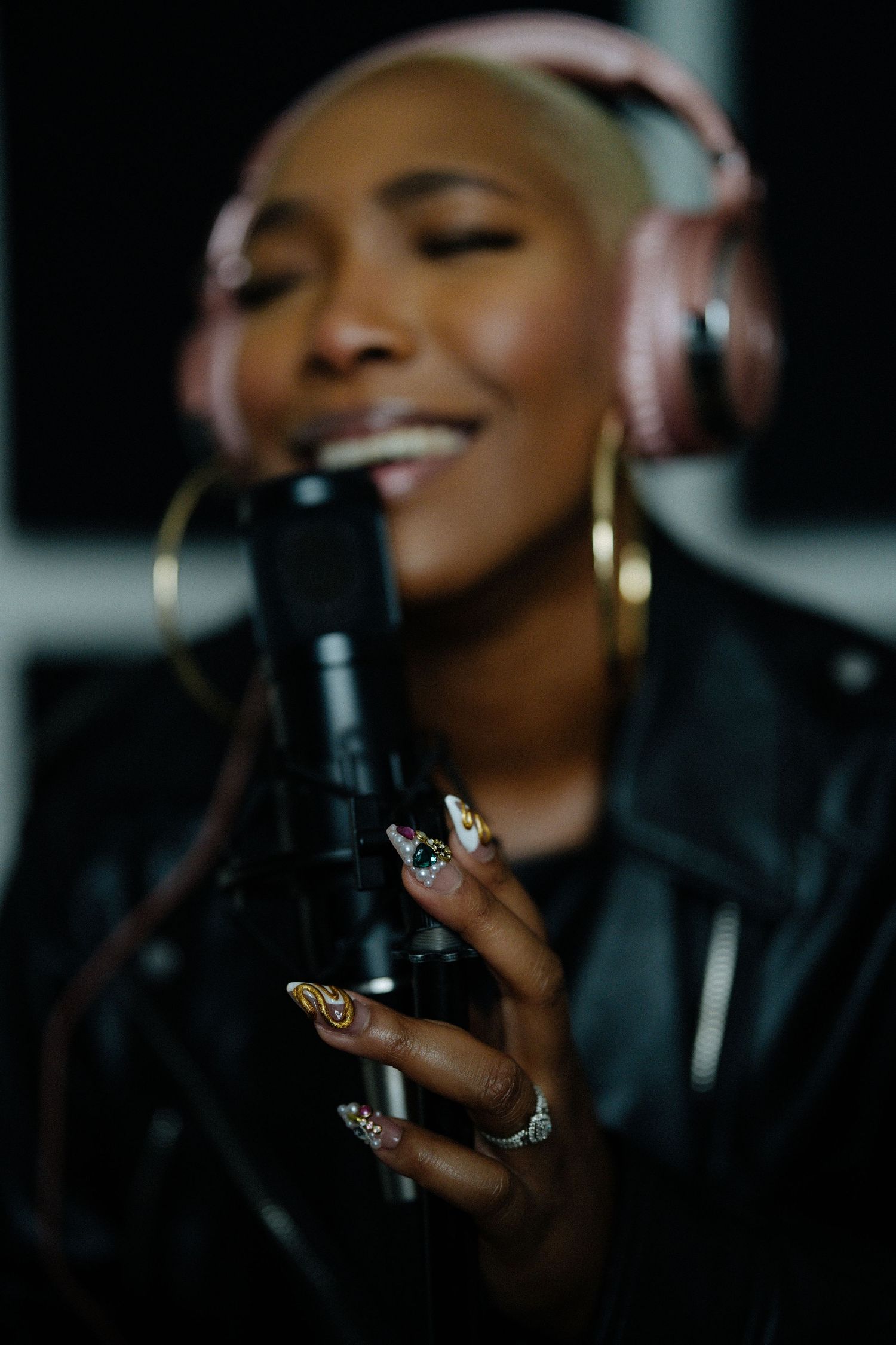 Close-up of decorated acrylic nails holding studio microphone with singer wearing pink headphones in background.