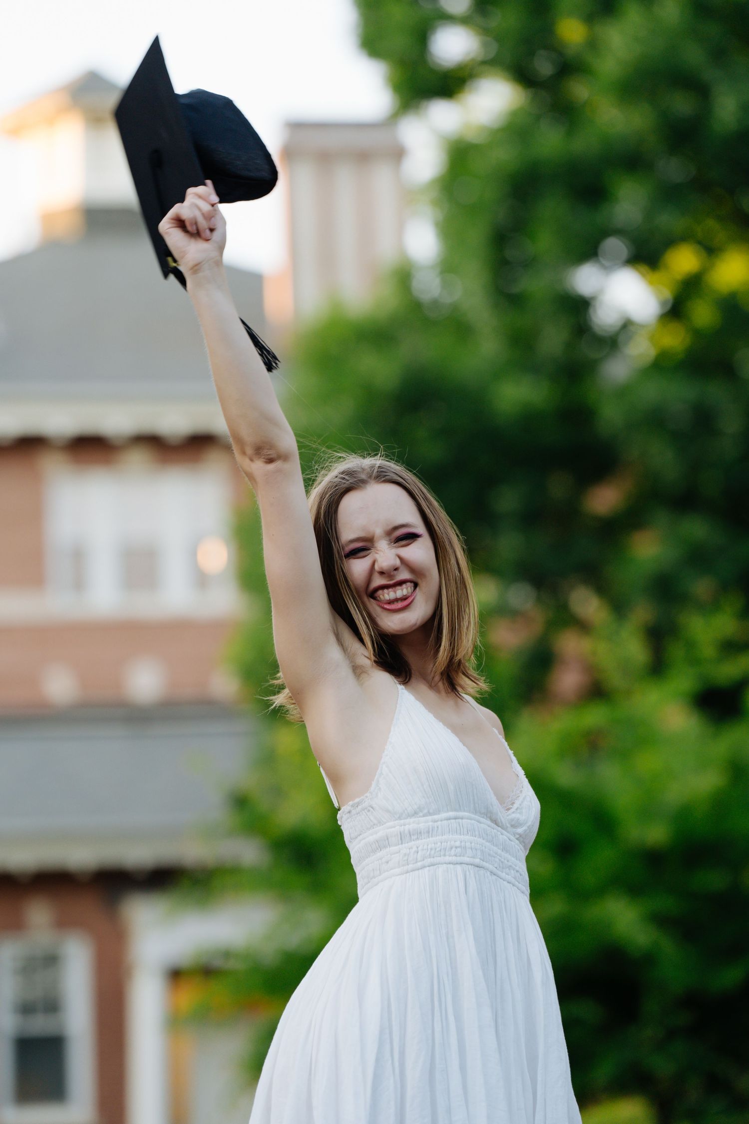 Excited female graduate in white dress raises cap triumphantly on campus.