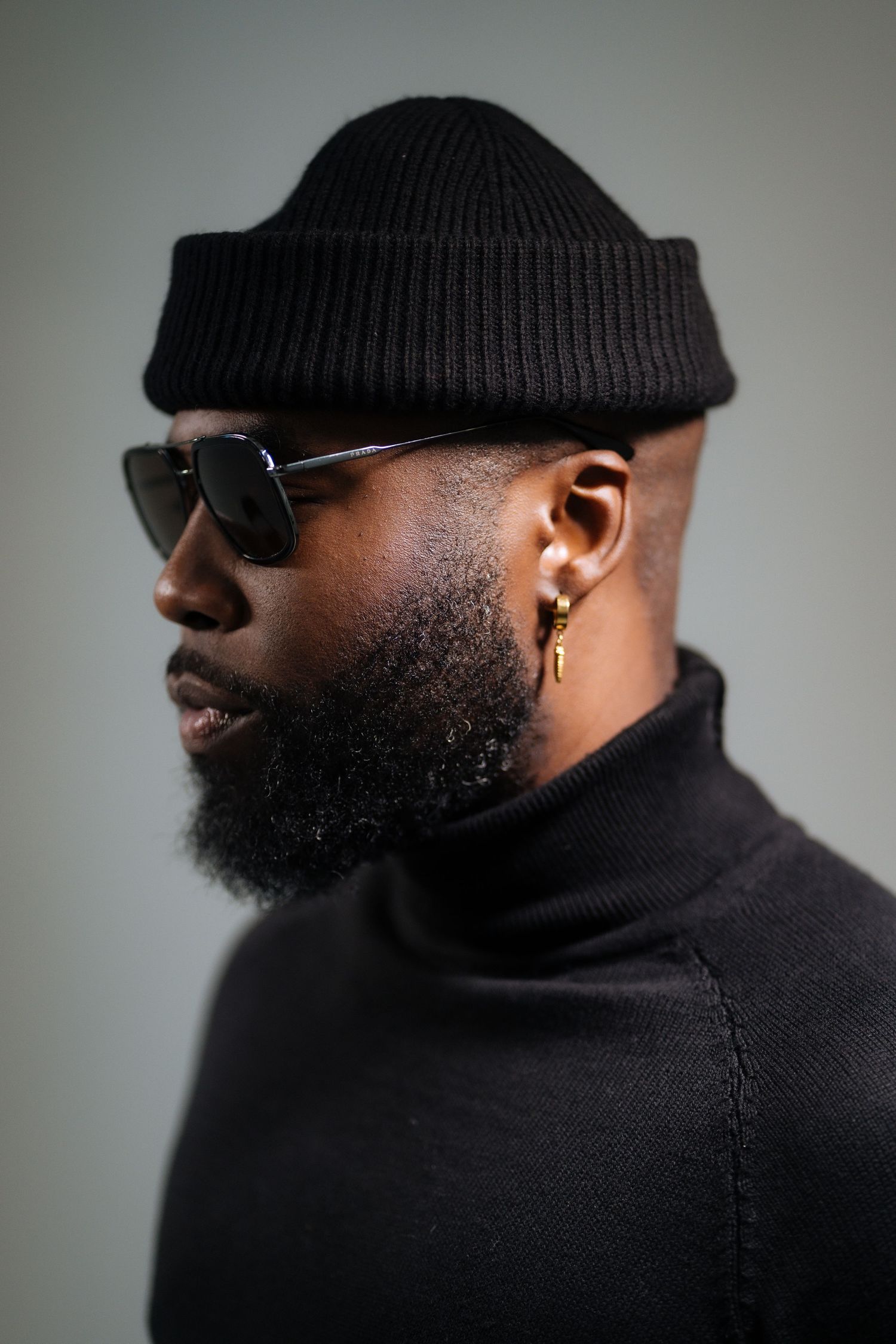 Side profile of a Black man with beard wearing black turtleneck, sunglasses, beanie, and gold earring.