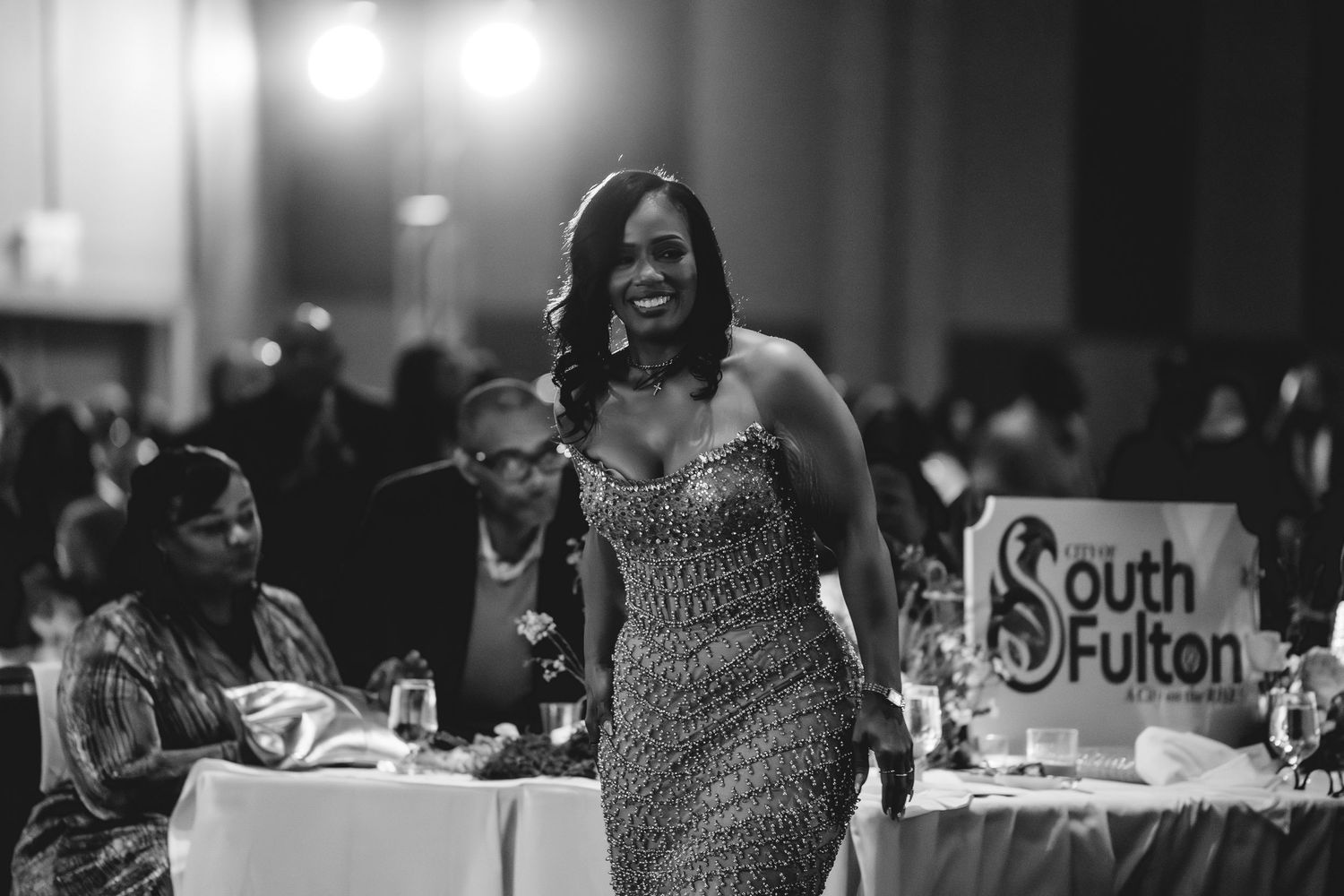 Elegantly dressed woman in sequined gown walking confidently at a South Fulton event in black and white.