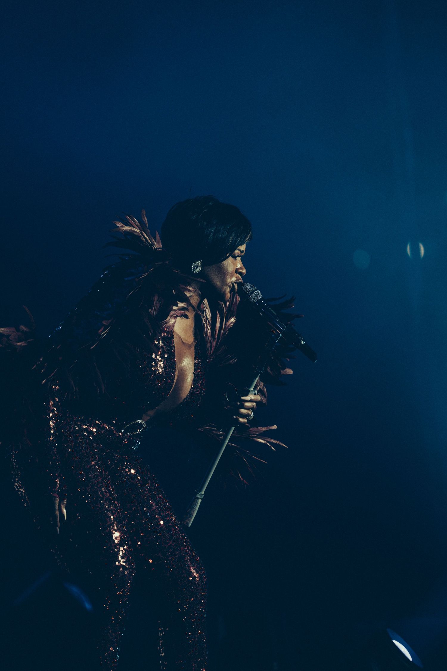 Female singer in feathered costume performing on stage under dramatic blue spotlight.