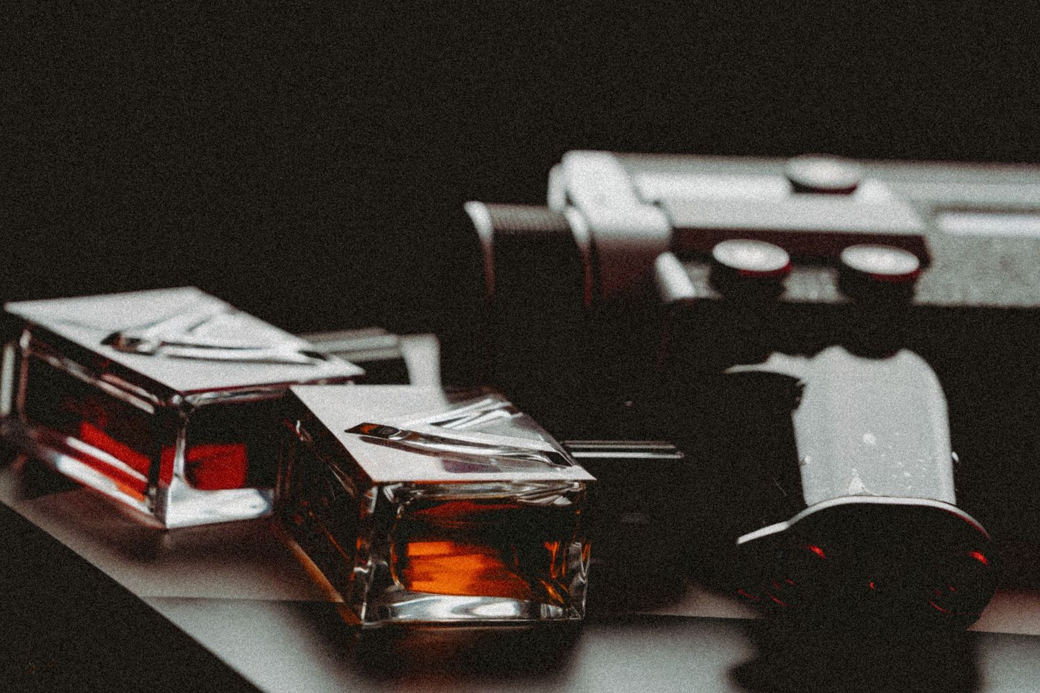 Two small glass perfume bottles with amber liquid sit on a dark table beside a blurred black camera in moody, low-key lighting.
