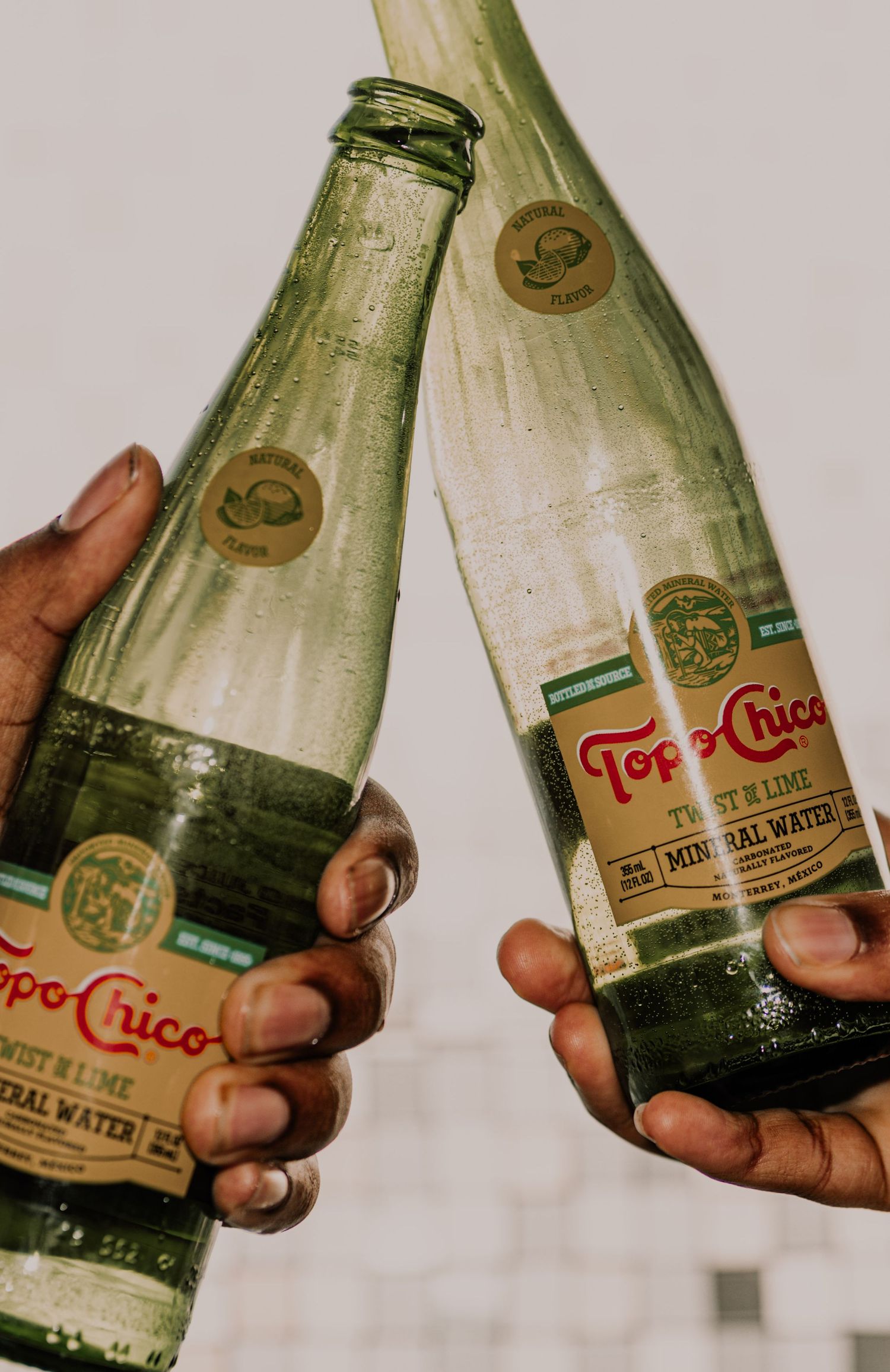 Two hands clinking Topo Chico Twist of Lime mineral water glass bottles together.