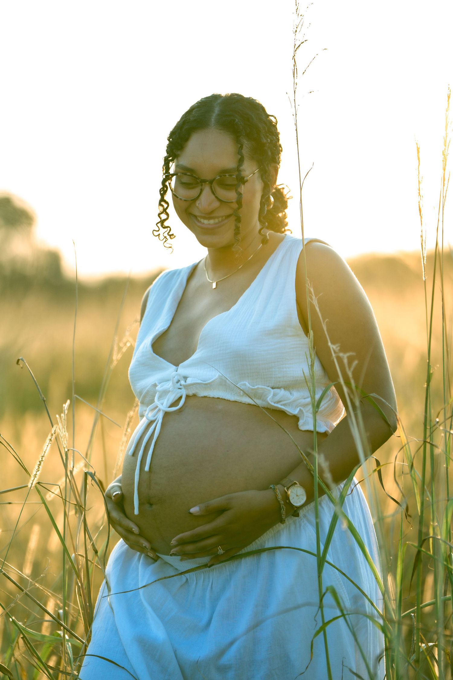 Happy pregnant woman in white dress cradles baby bump in sunlit grass field.