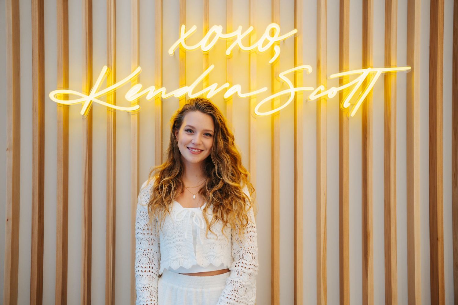 Woman with curly hair in white crop top posing by XOXO Kendra Scott neon sign.