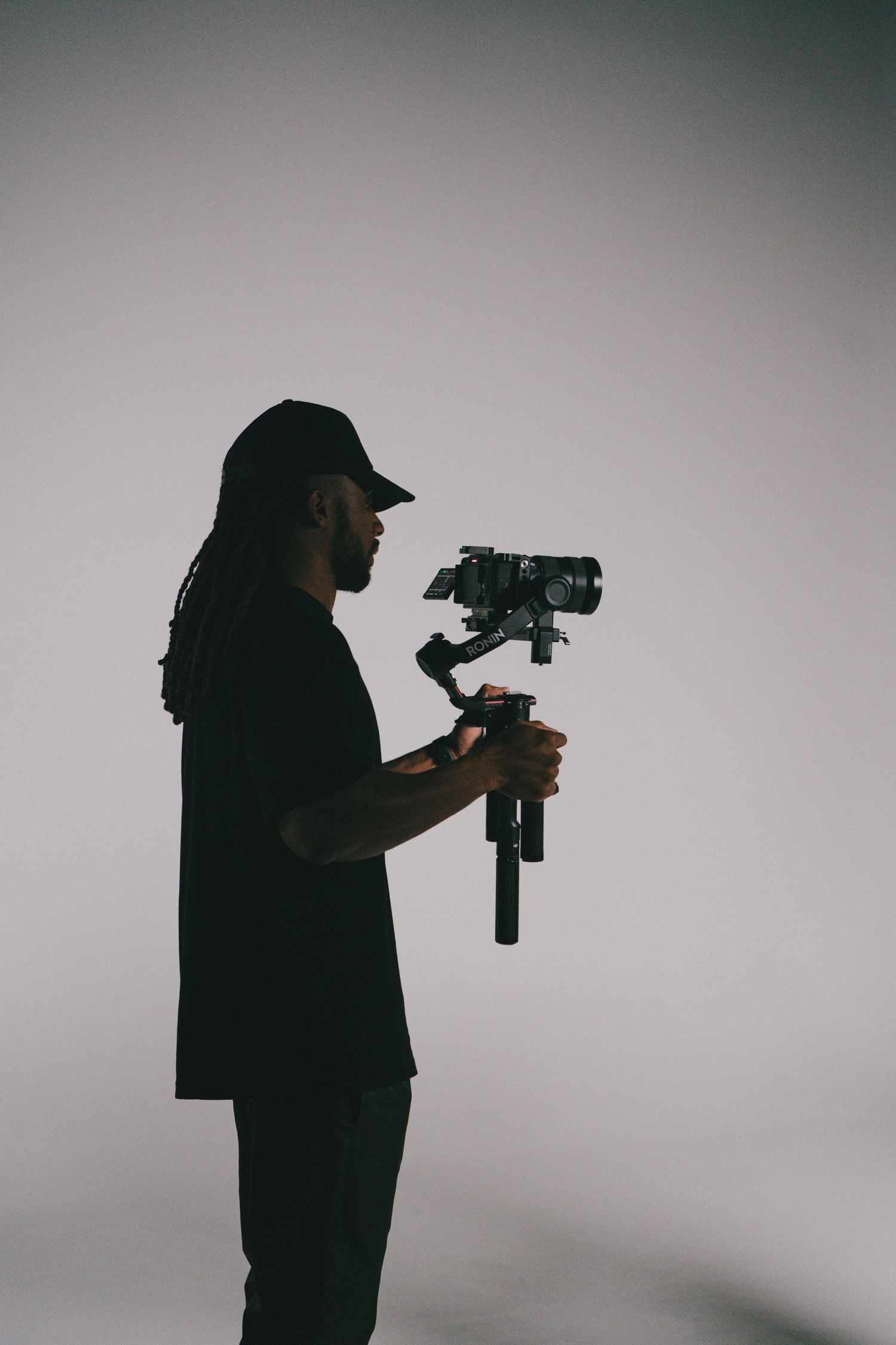 Silhouette of a videographer with dreadlocks holding a camera gimbal stabilizer against a white studio background.