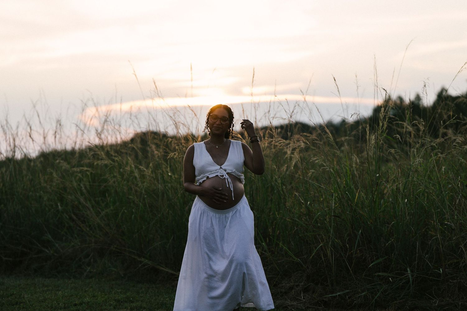 Pregnant woman in white dress holds belly during stunning sunset outdoors.