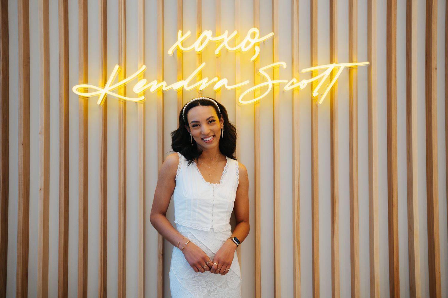 Smiling woman in white dress posing in front of XOXO Kendra Scott neon sign.
