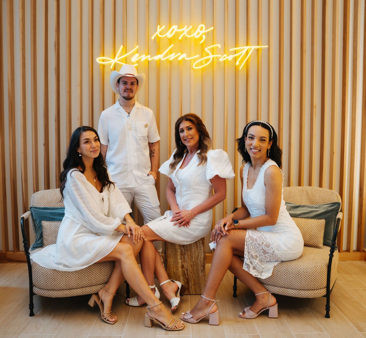 Four people in white outfits posing in front of XOXO Kendra Scott neon sign.