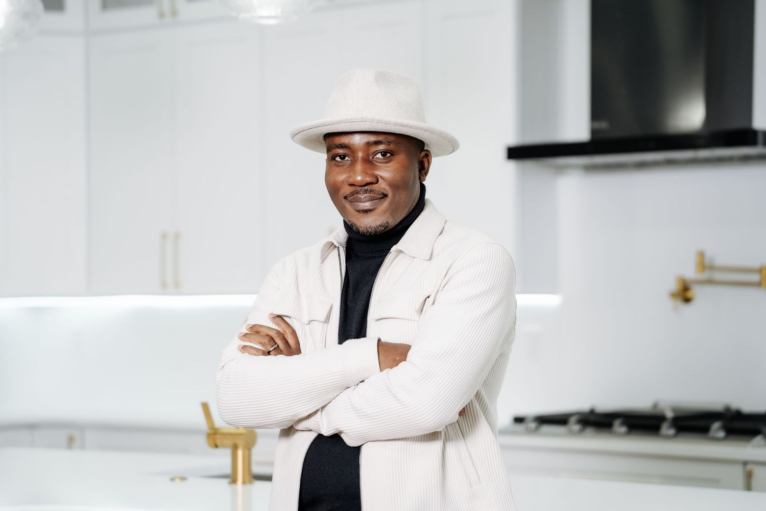 Confident Black man in white hat and jacket posing with arms crossed in kitchen.