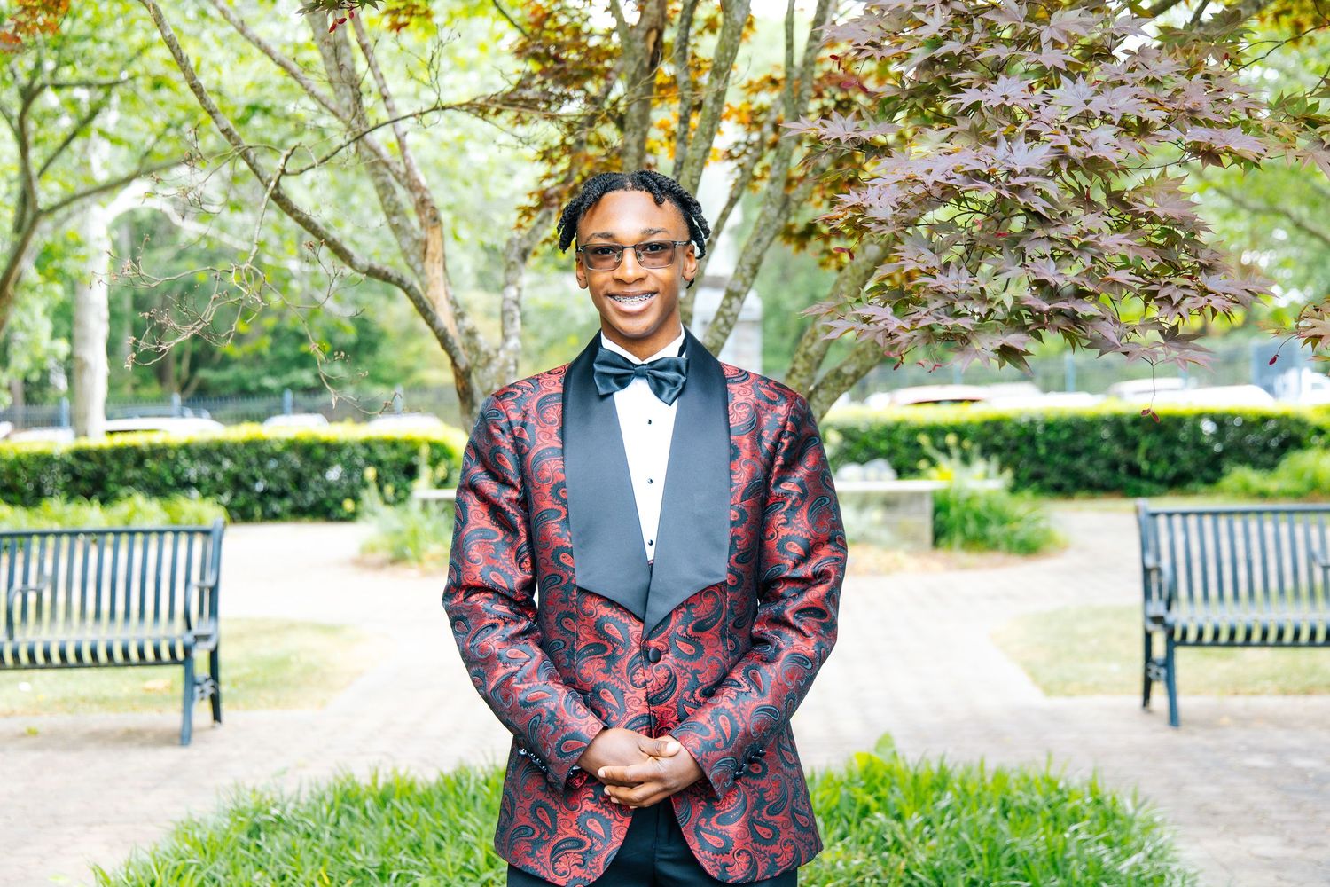 Young man in burgundy paisley tuxedo jacket with bow tie posing in park setting.