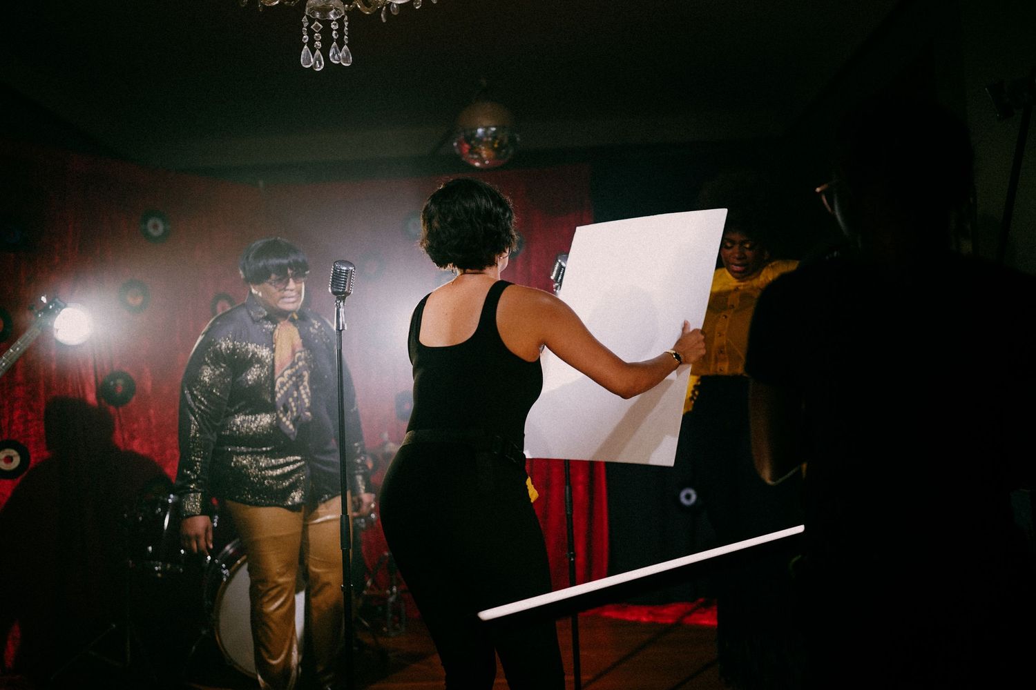 Film crew members work on a dimly lit music video set with red curtains and stage lighting.