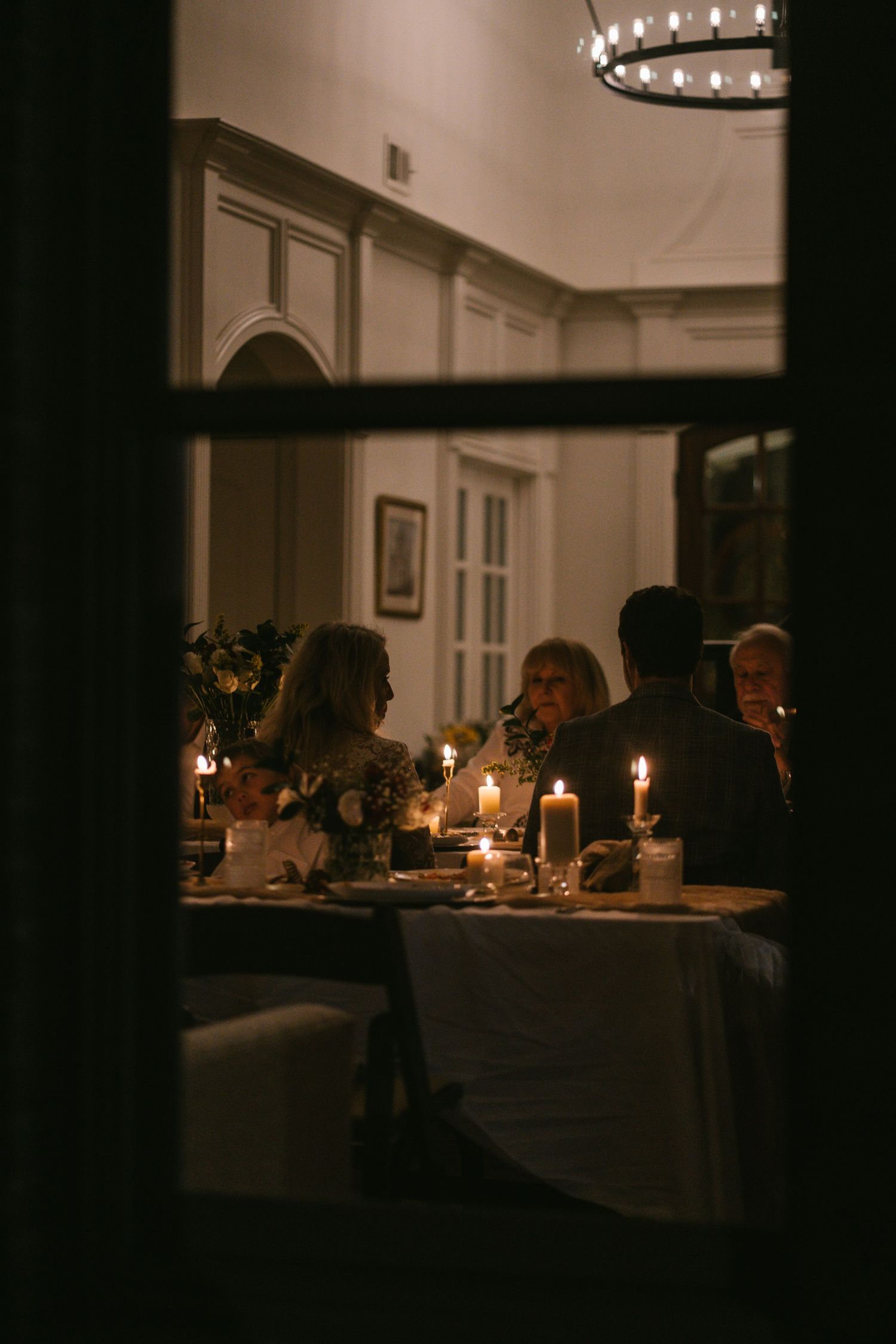 Guests dining by candlelight at an elegant dinner party viewed through a window.