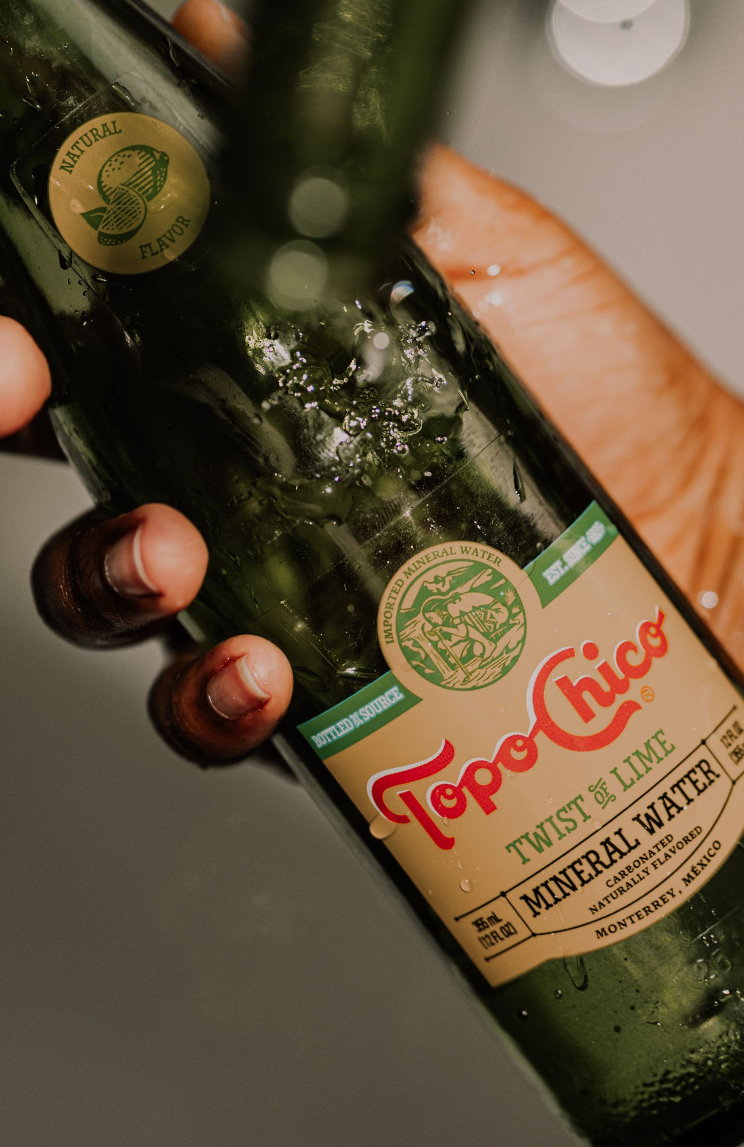 Hand holding a wet Topo Chico Twist of Lime Mineral Water green glass bottle.