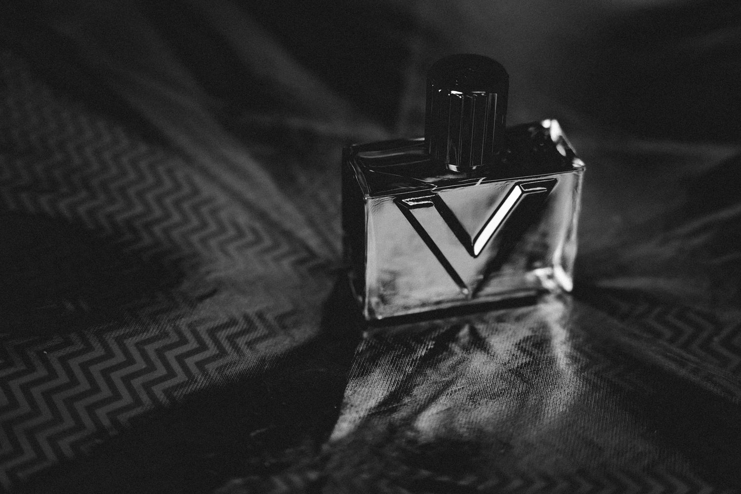 Black and white photo of a rectangular perfume bottle with a bold V design and dark cap on a shiny zigzag patterned fabric.