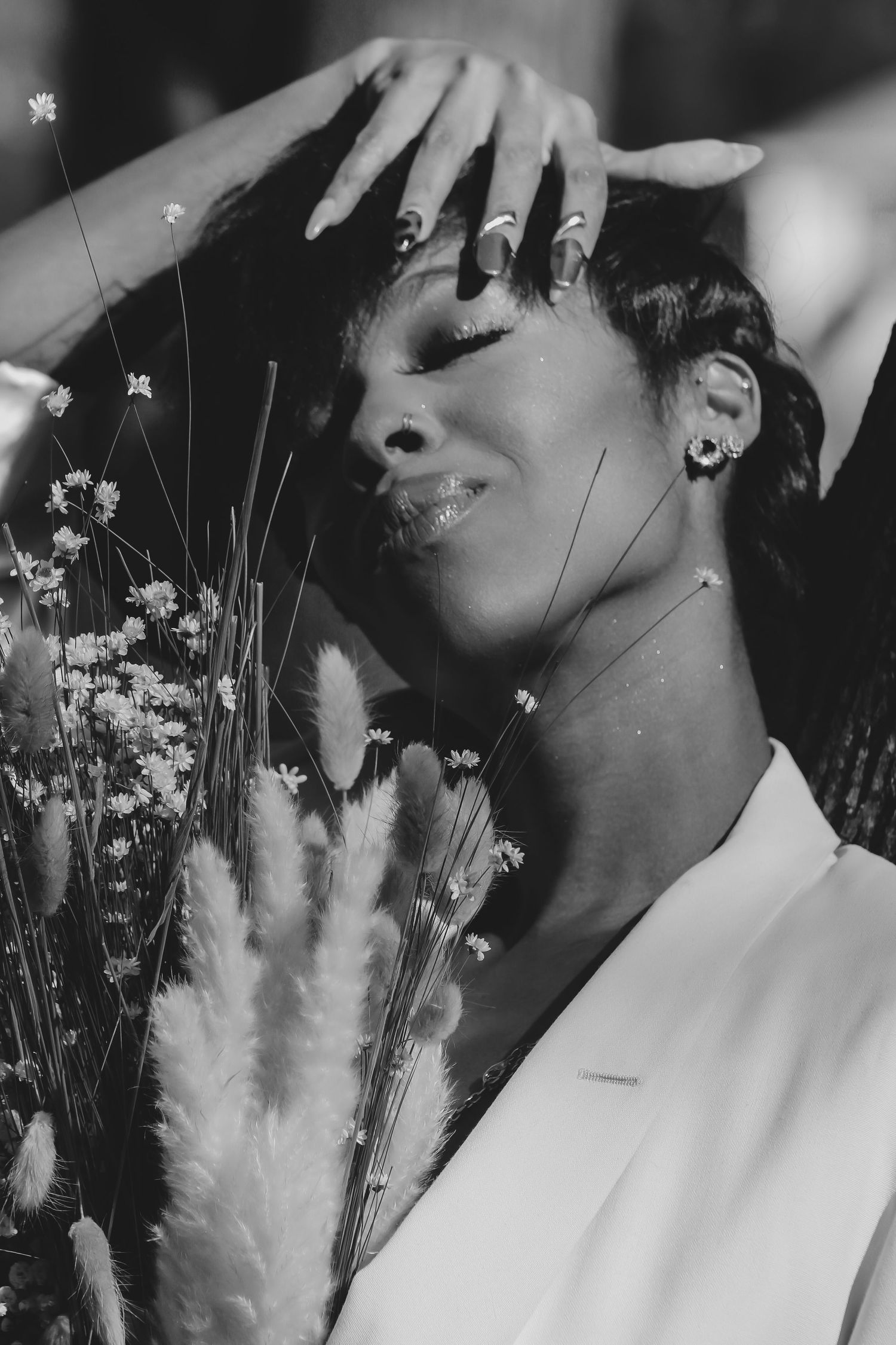 Black and white portrait of a woman in white blazer holding wildflowers, eyes closed, hand on head.