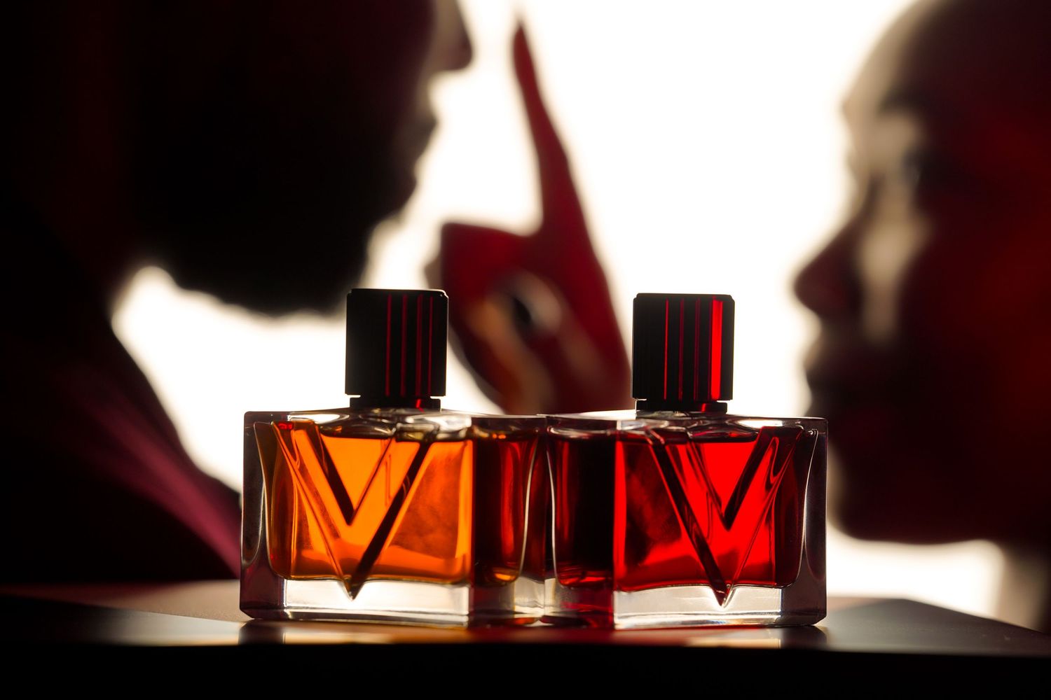 Two amber and red perfume bottles with V-shaped designs in front of two silhouetted faces in profile against a bright backdrop.