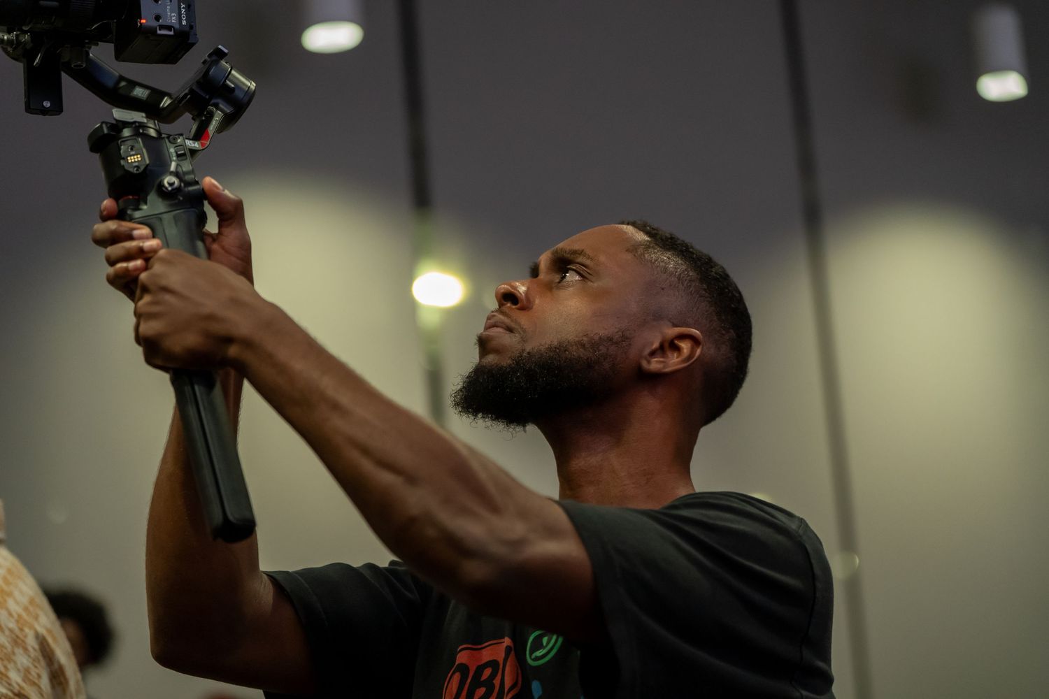 Videographer adjusts gimbal camera stabilizer at indoor event.