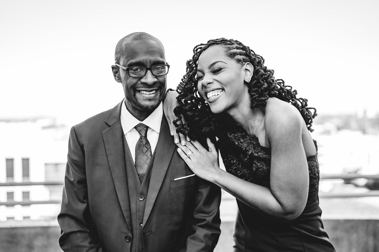 Black and white photo of a laughing couple in formal wear sharing a joyful moment on a rooftop.