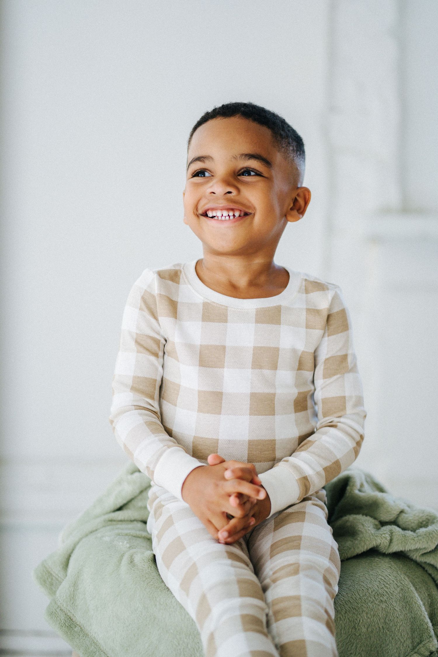 Adorable young Black boy in tan plaid pajamas sitting and smiling brightly.