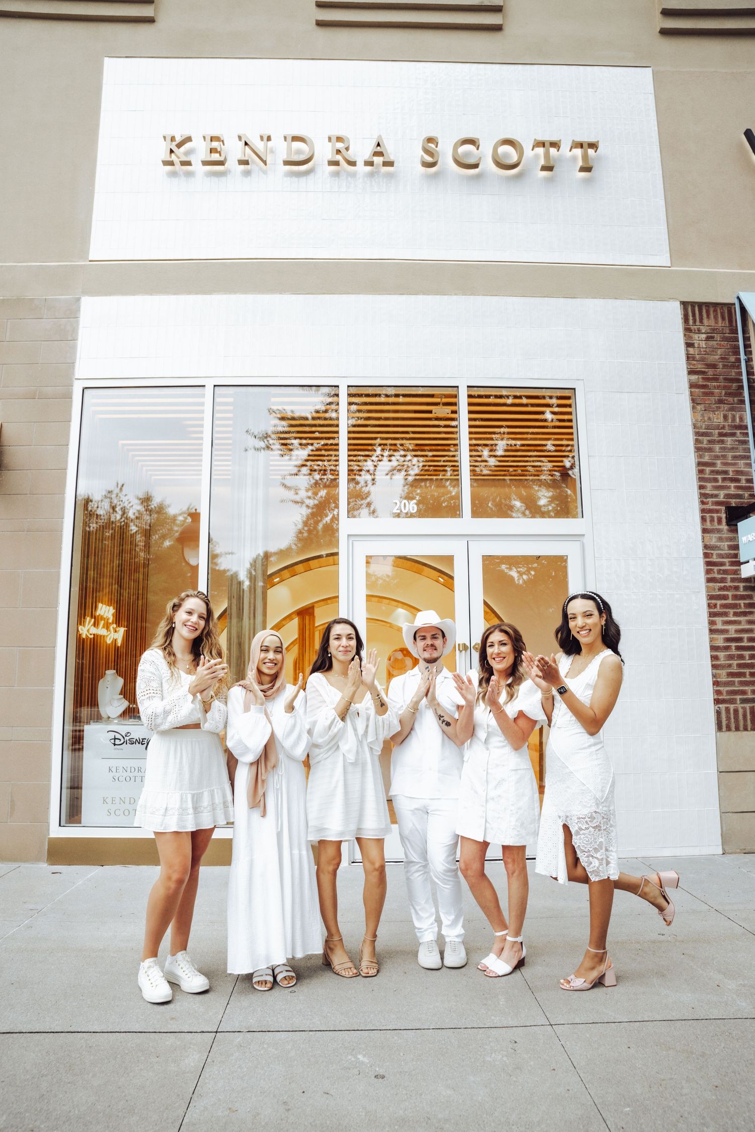 Six people in white outfits pose and clap in front of a Kendra Scott store entrance.