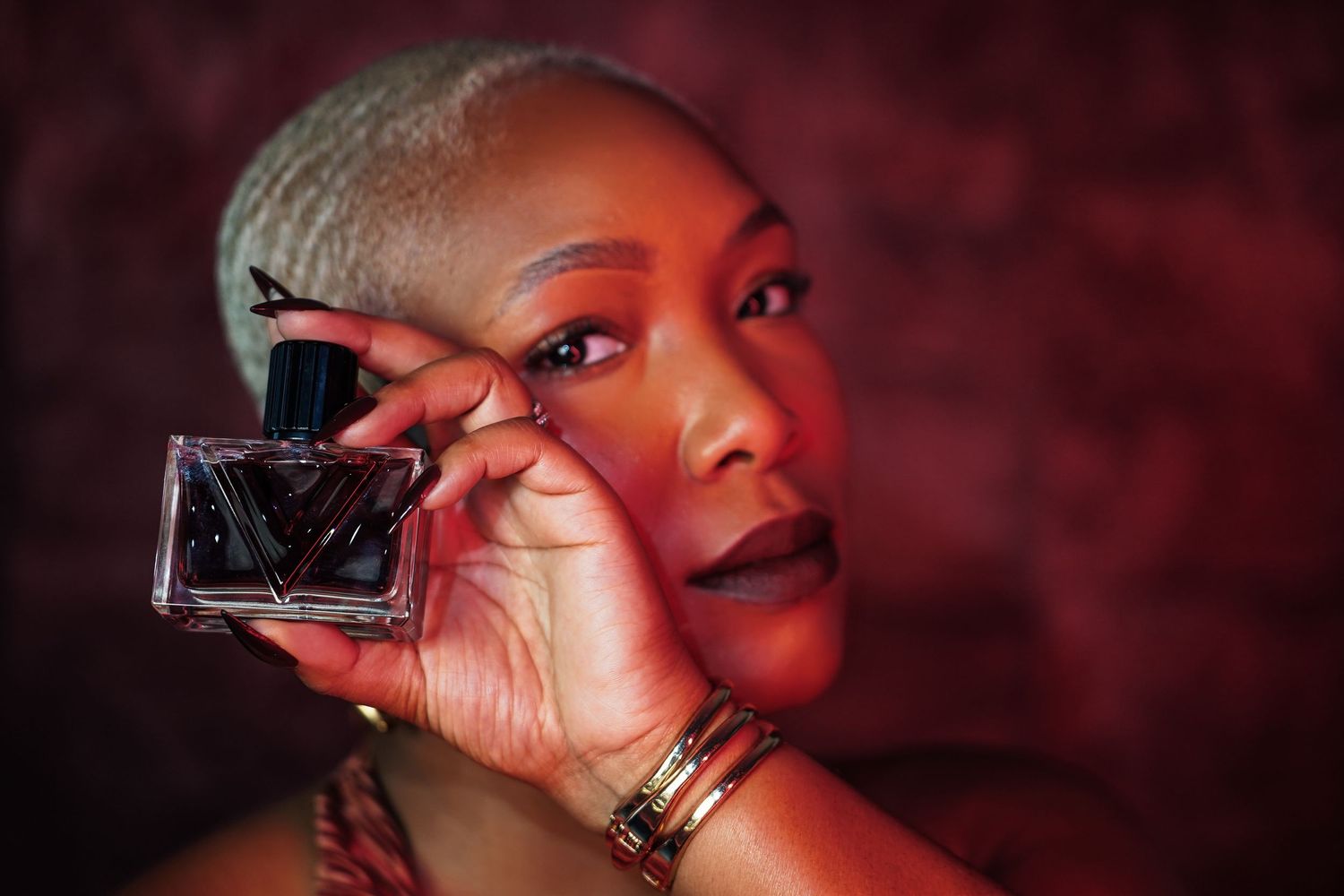 Woman with short silver hair and gold bangles holds a clear perfume bottle beside her face against a moody red background.