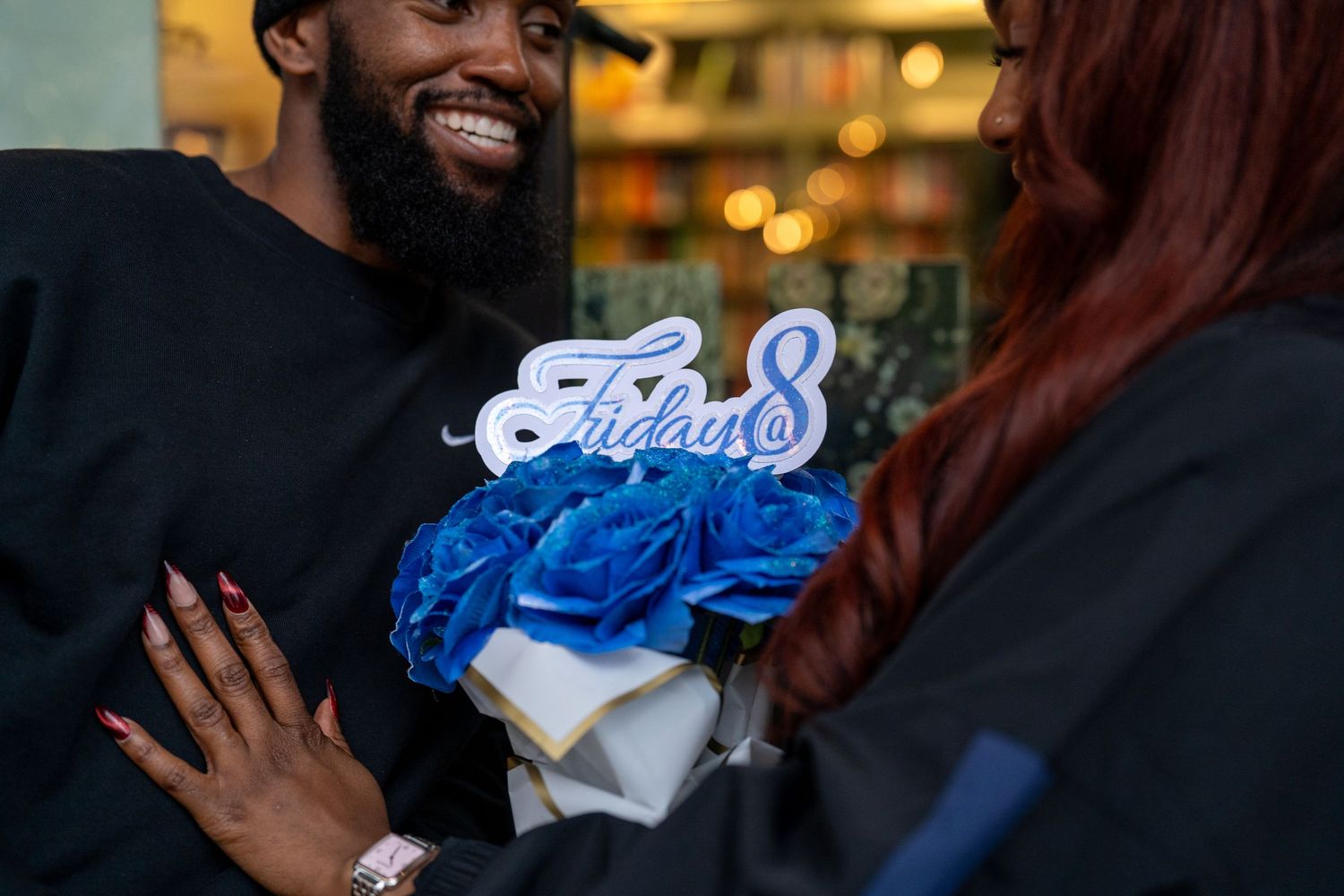 Man presenting blue roses bouquet with Friday@8 sign to woman in black outfit.