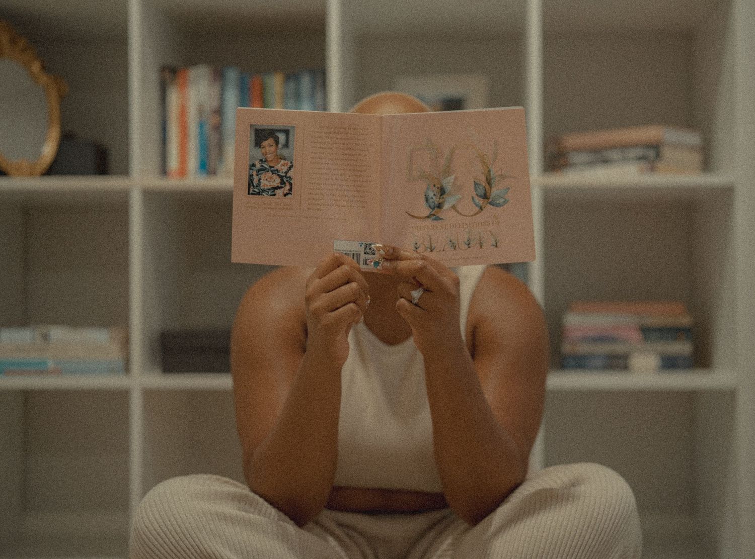 Person sitting cross-legged holding open colorful book in front of face near bookshelf.