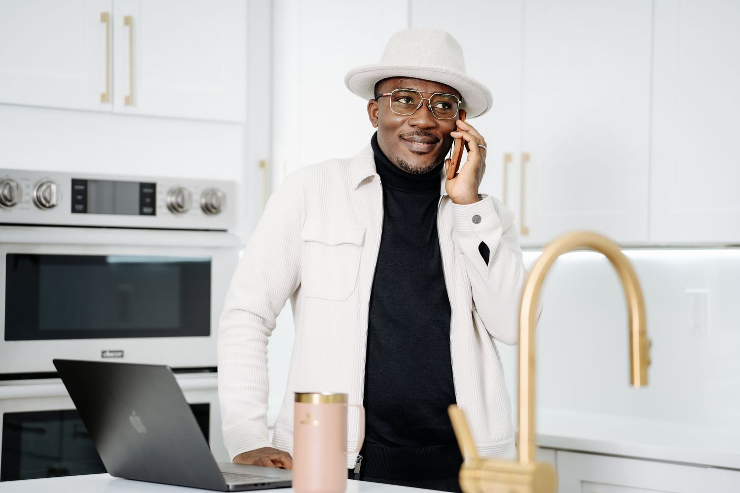 Man in white hat talking on phone by laptop in modern white kitchen.