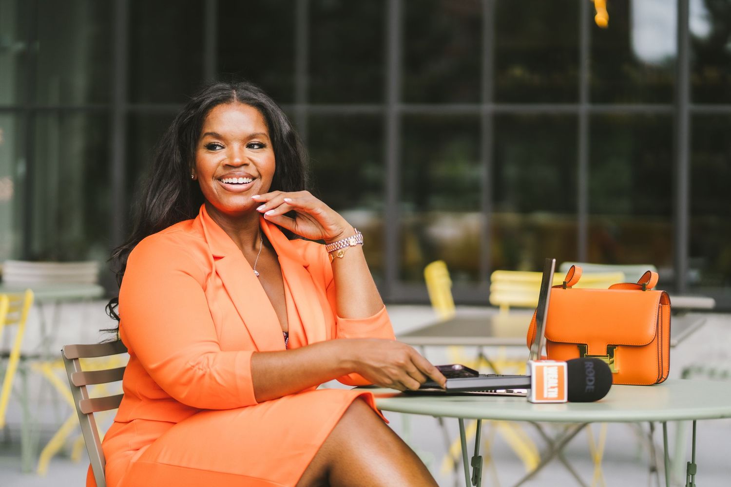 Smiling woman in orange blazer sitting at outdoor table with laptop, microphone, and orange handbag.