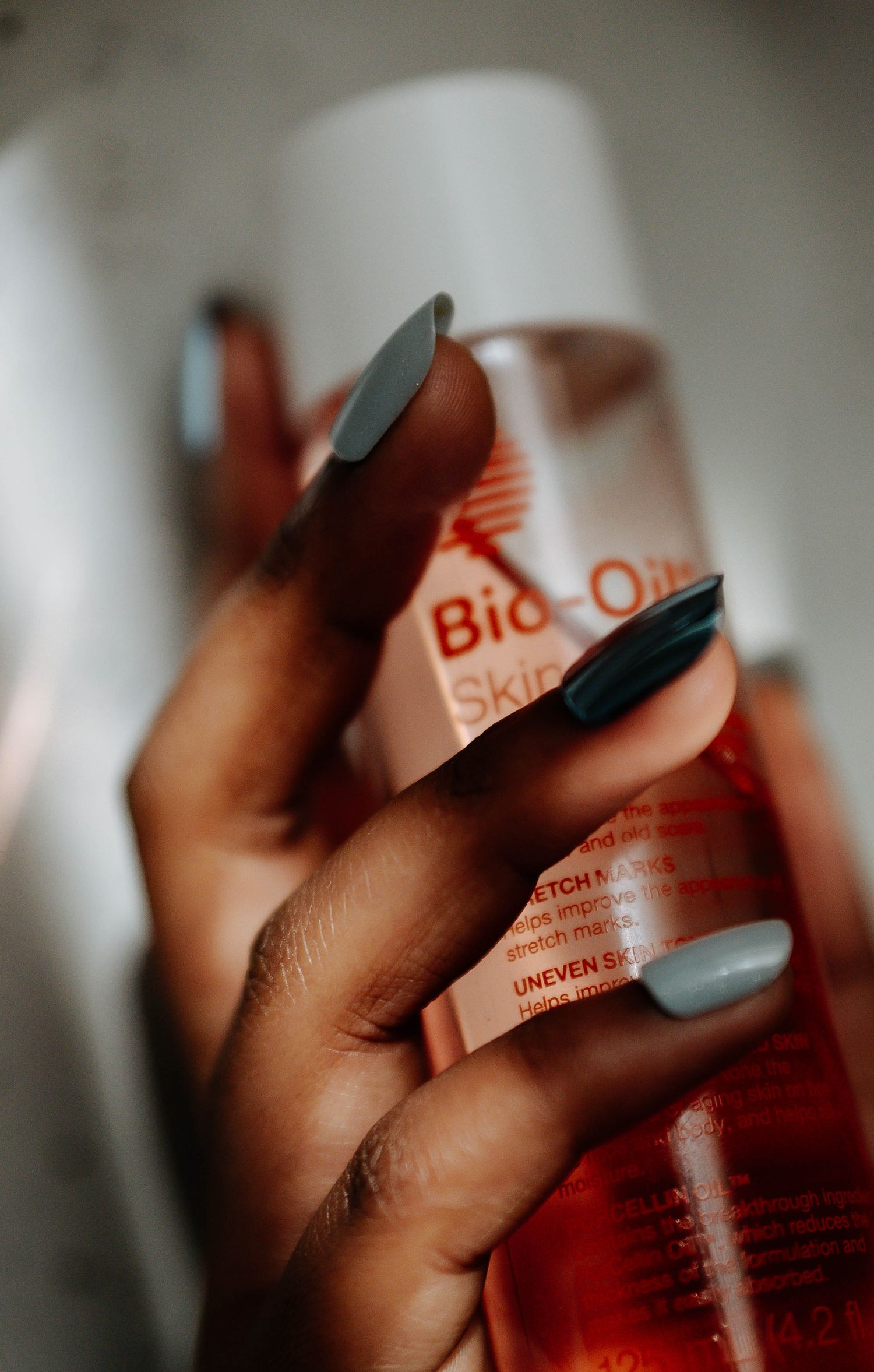 Close-up of hand with gray nails gripping a Bio-Oil Skincare Oil bottle in soft light.