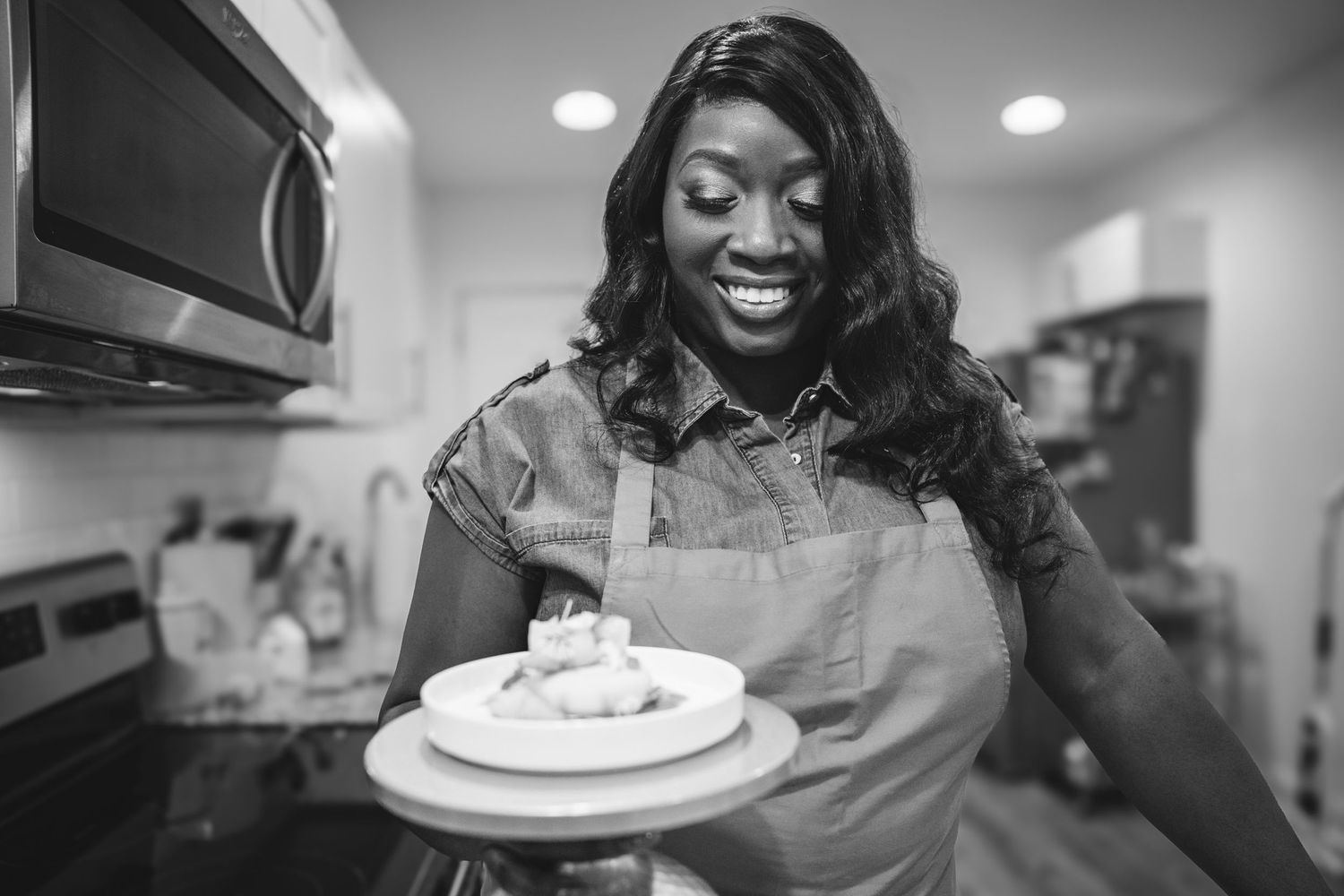 Happy Black woman chef in apron presenting a plate of food in kitchen, black and white.