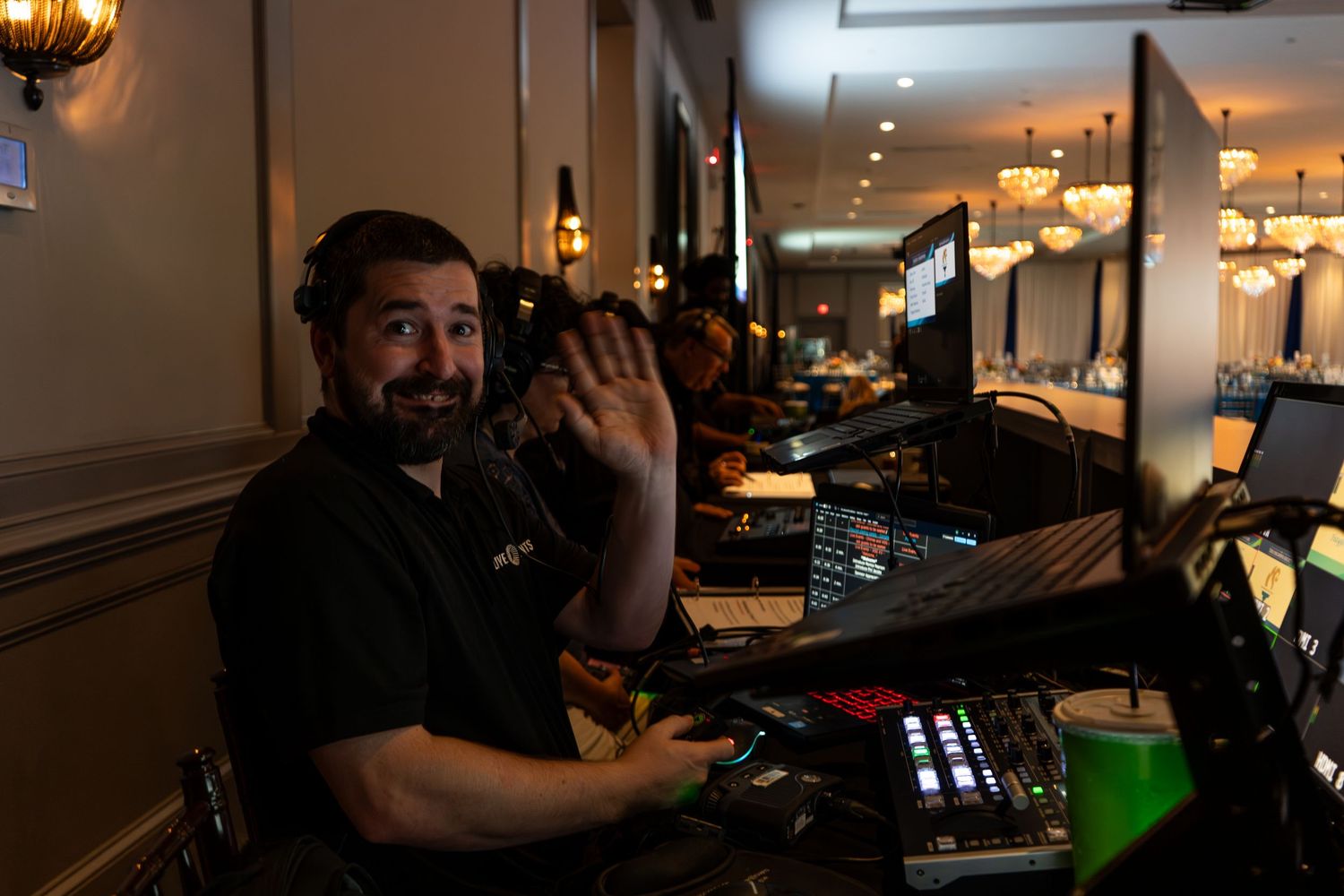 A smiling man waves at a camera while working at a professional audio-visual setup in an elegant ballroom venue.