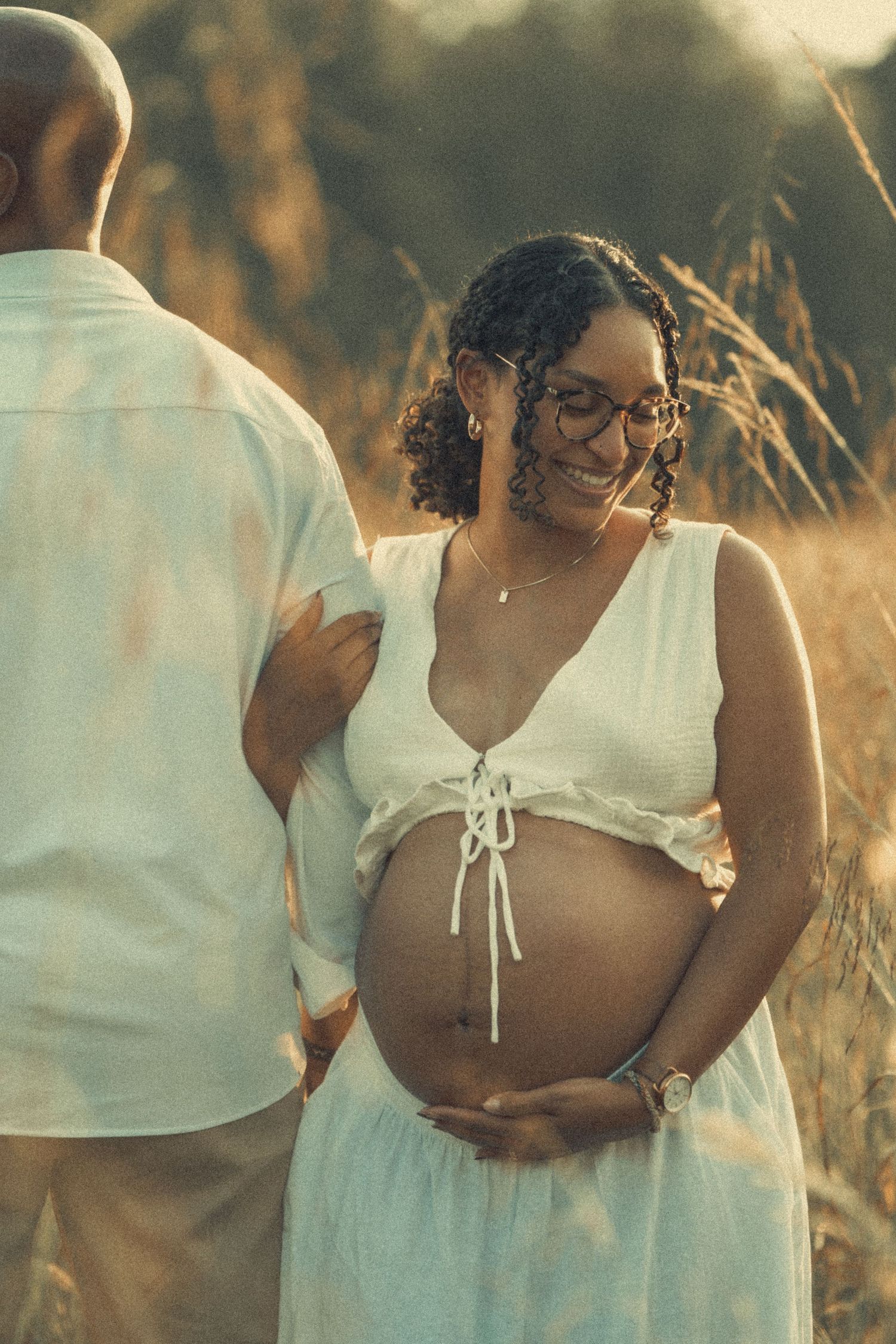 Smiling pregnant woman in white dress cradling her baby bump in a sun-drenched golden field.