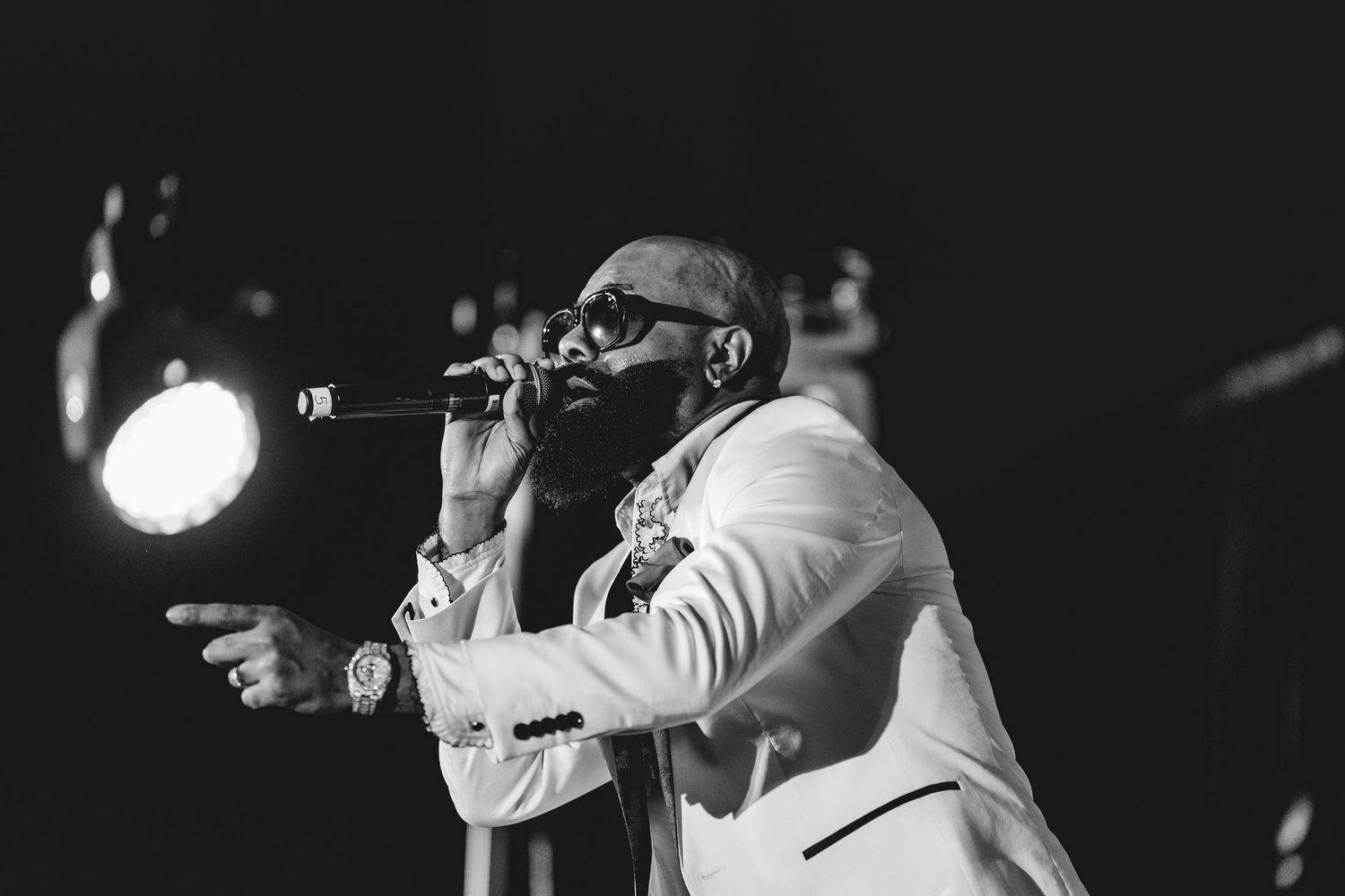 Bearded male performer in white suit singing passionately into microphone on a dimly lit stage.