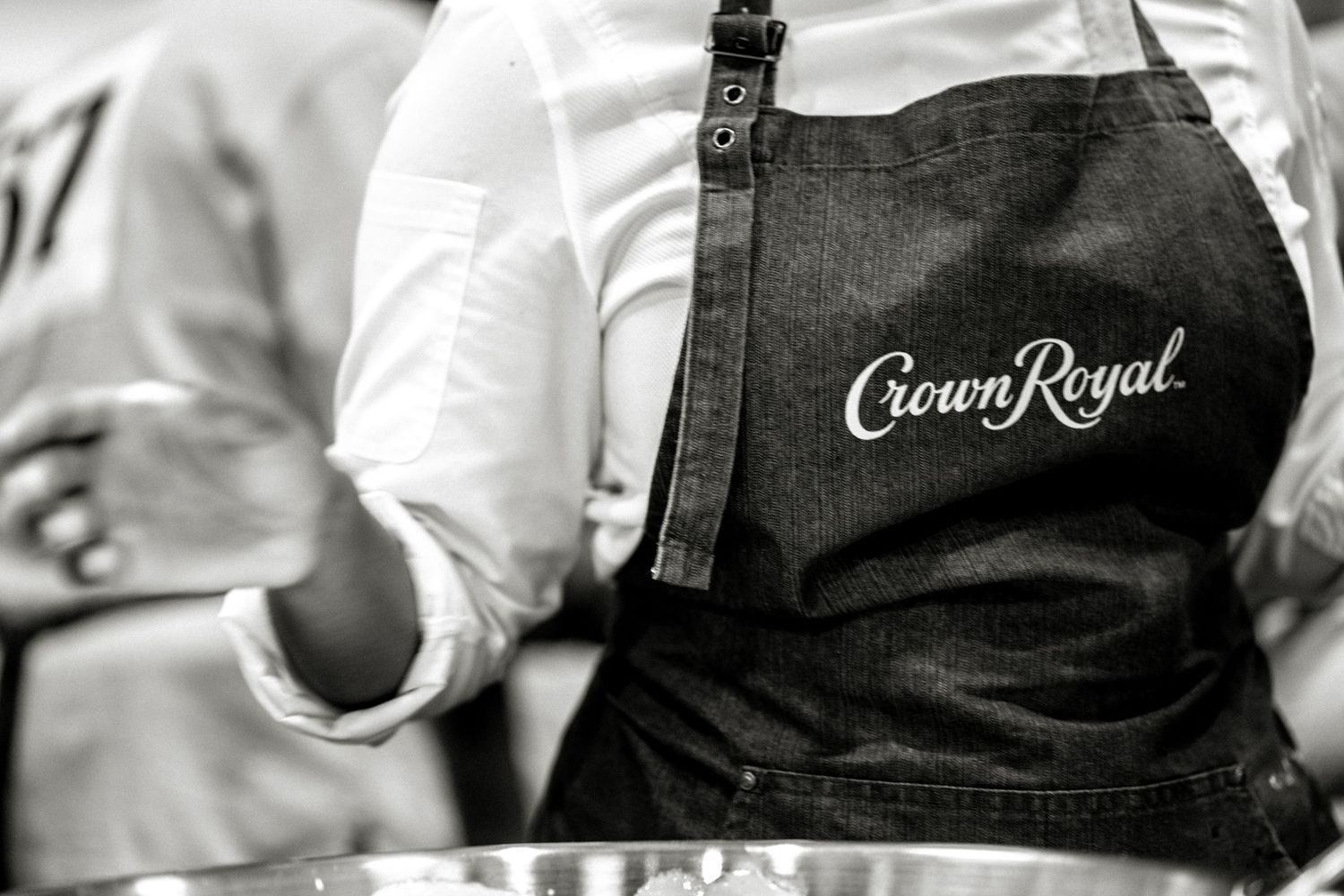 A person wearing a black Crown Royal branded denim apron stands at a culinary station.