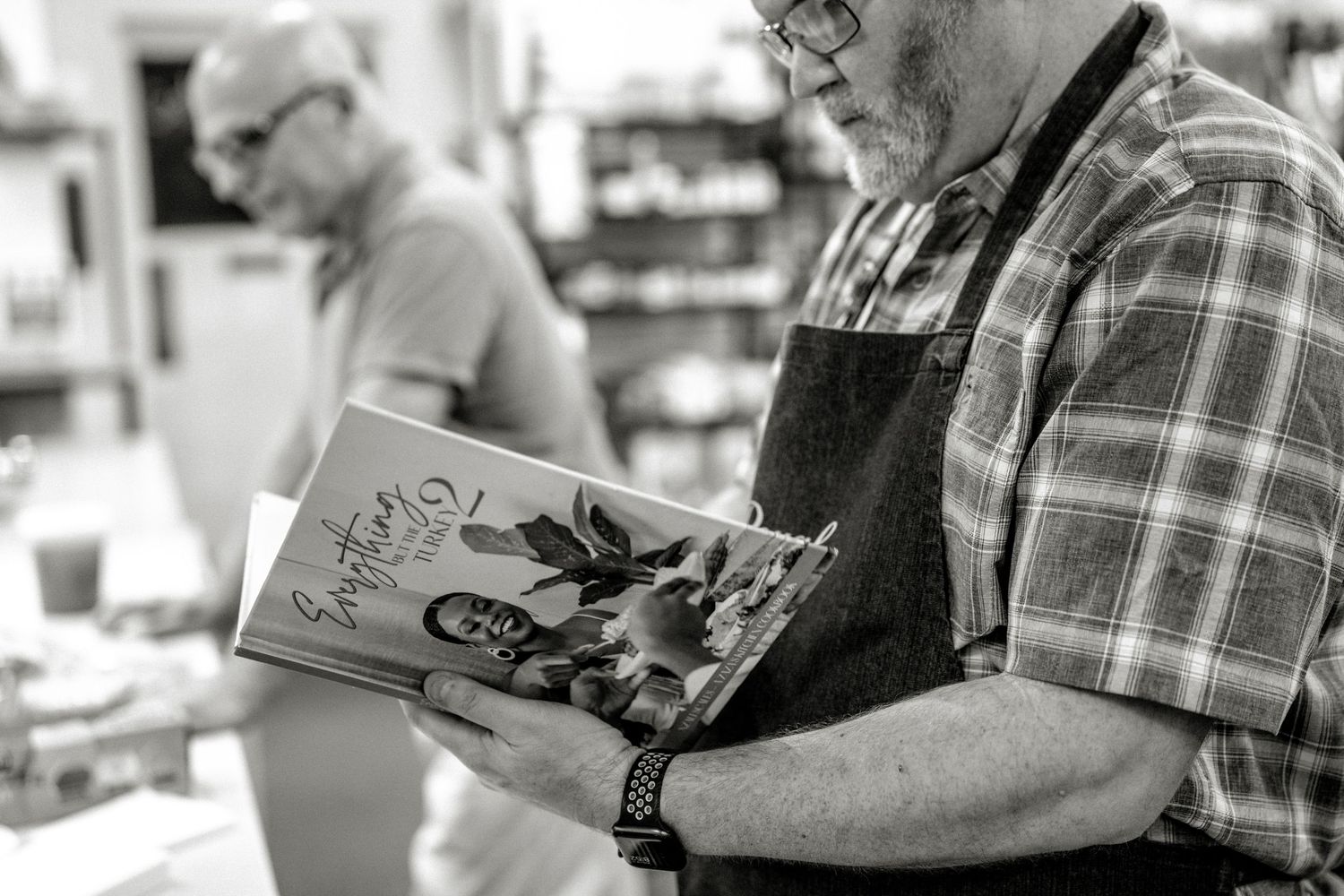 Man in plaid shirt and apron reads a cookbook in a store.